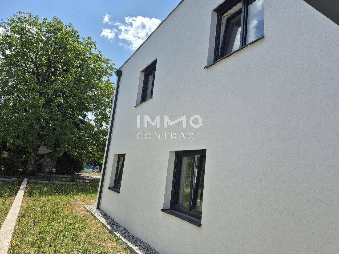 Moderne weiße Fassade eines Neubaus mit dunklen Fensterrahmen und umliegender Grünfläche.