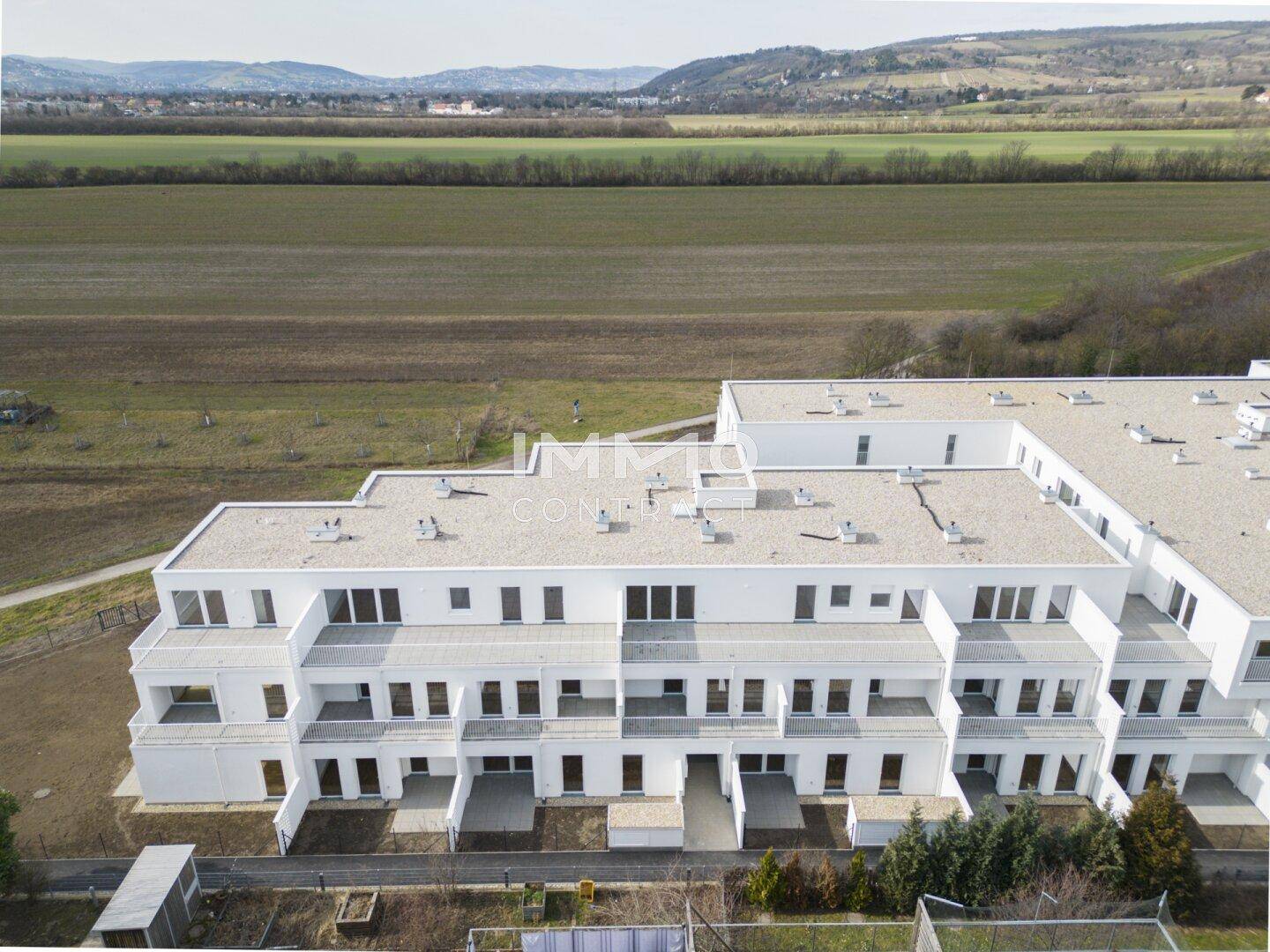 Moderne weiße Wohnanlage mit Flachdächern und Balkonen, umgeben von grünen Feldern und Hügeln.