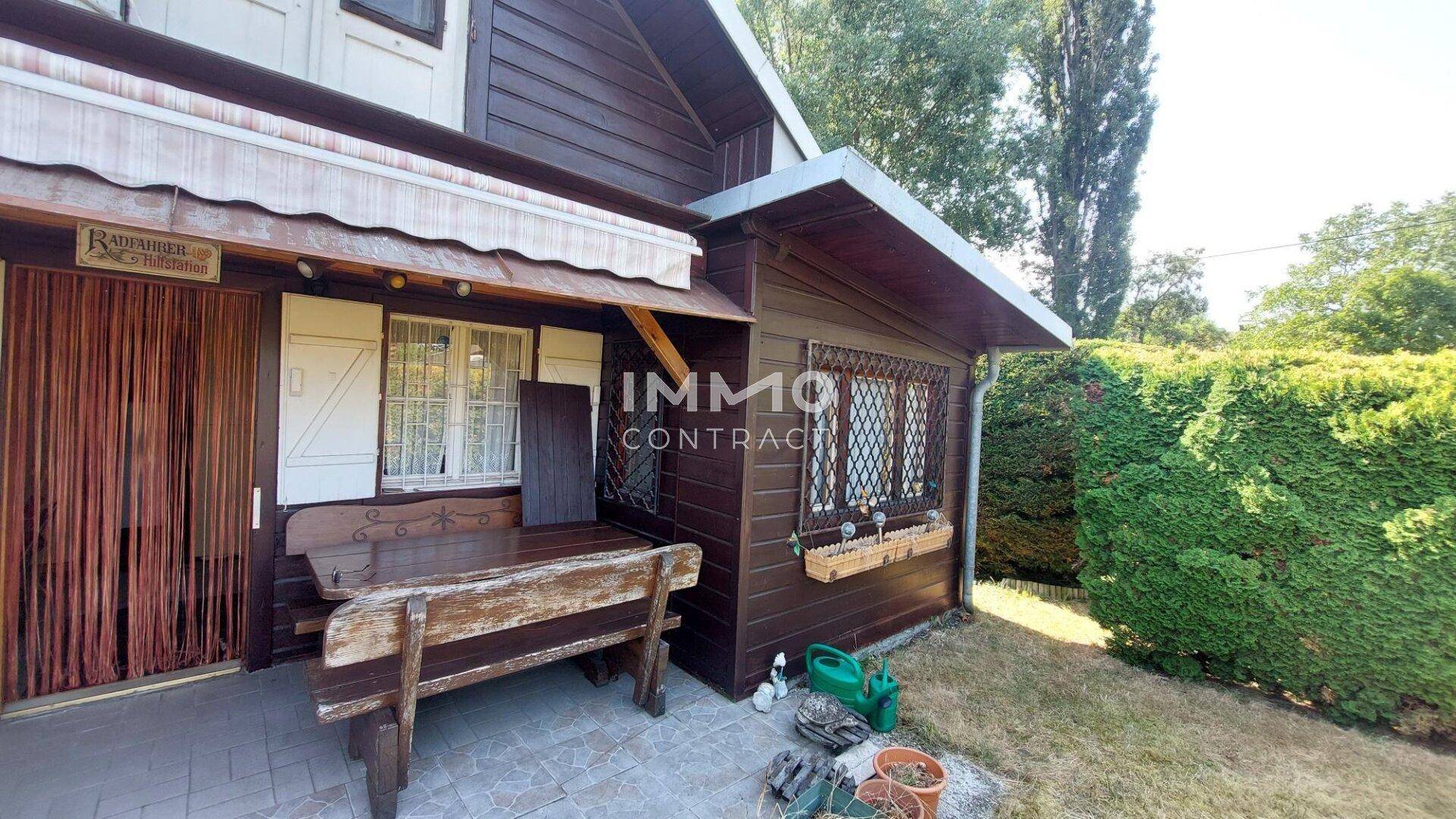 Rustikales Holzhaus mit kleiner gepflasterter Terrasse und Holzsitzgelegenheiten im Grünen.