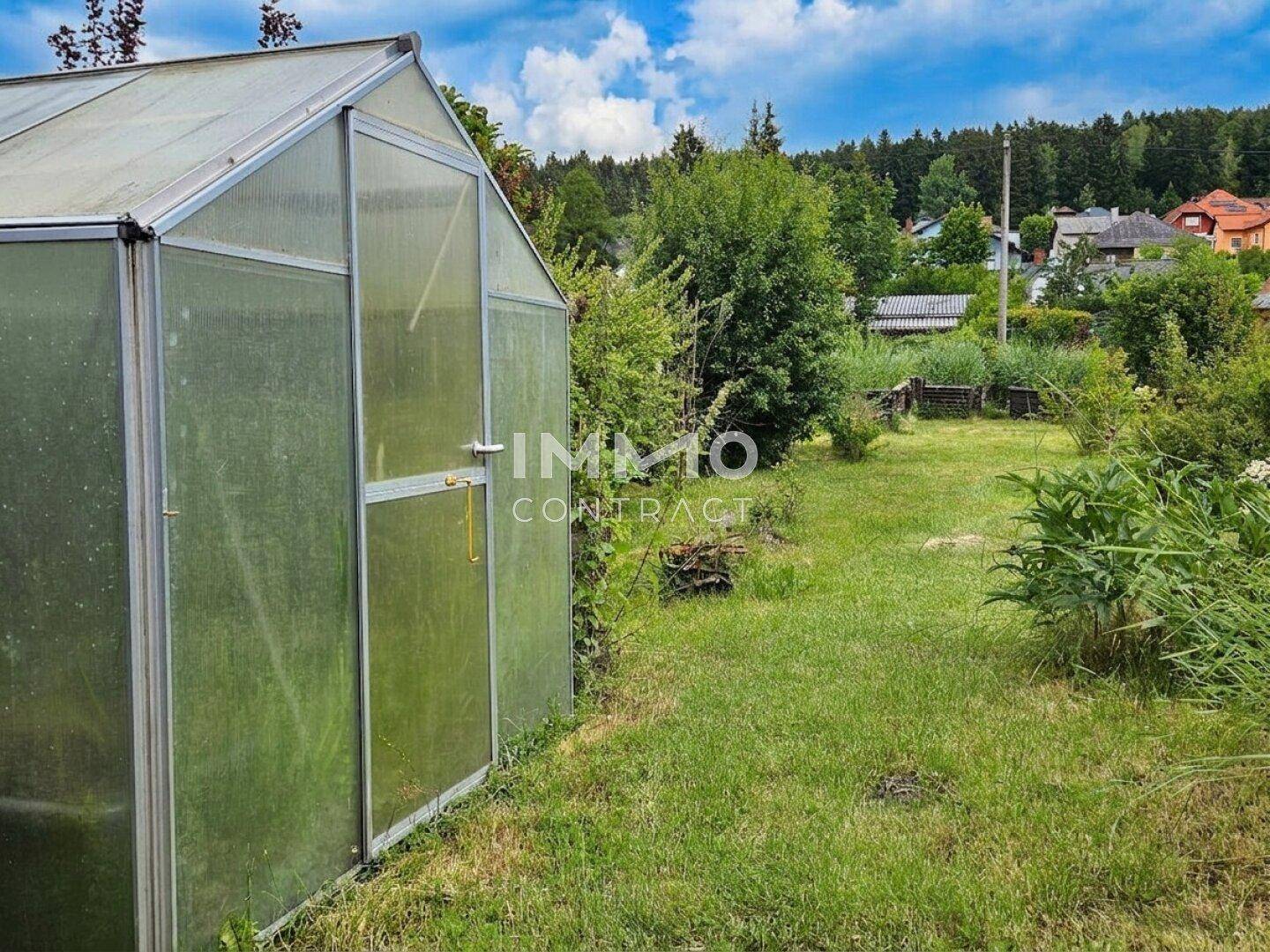 Gartenbereich mit einem Gewächshaus und Blick auf die bewaldete Umgebung.