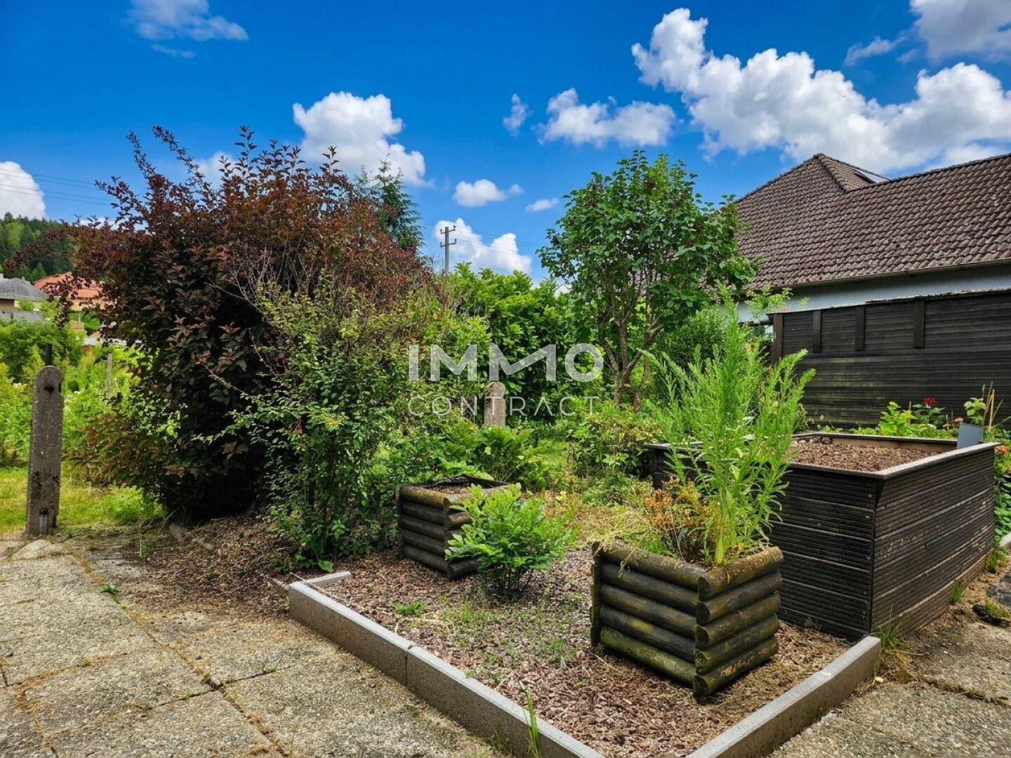 Gartenweg mit Hochbeeten und Sträuchern, im Hintergrund ein Teil des Hauses.