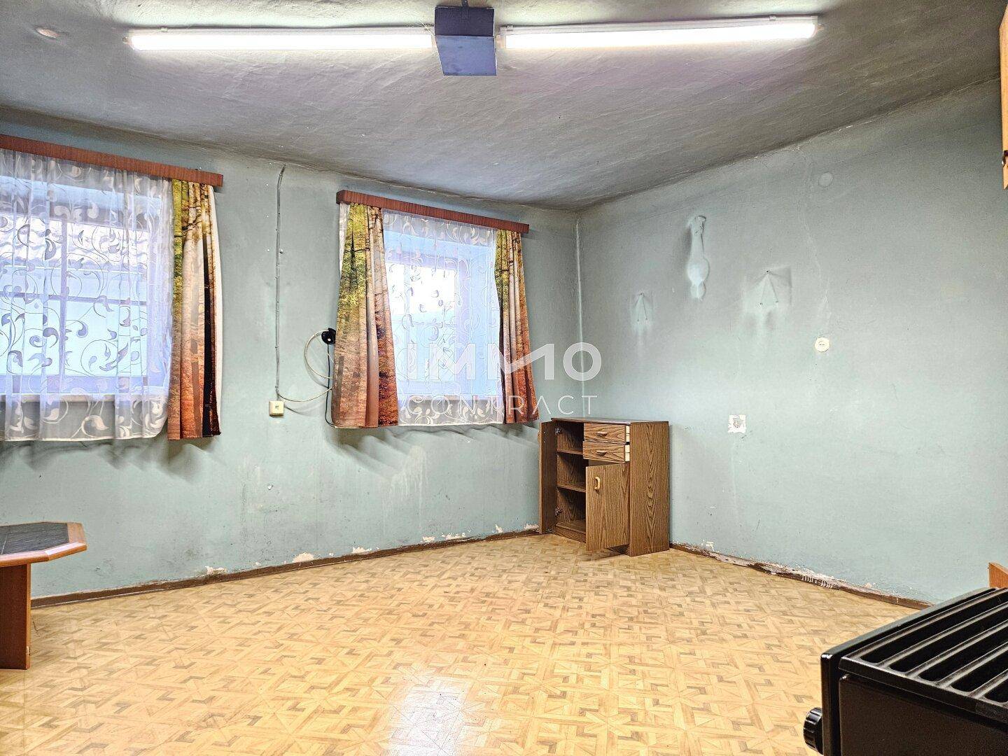 Wohnraum mit zwei Fenstern, Linoleumboden und hellblauen Wänden.