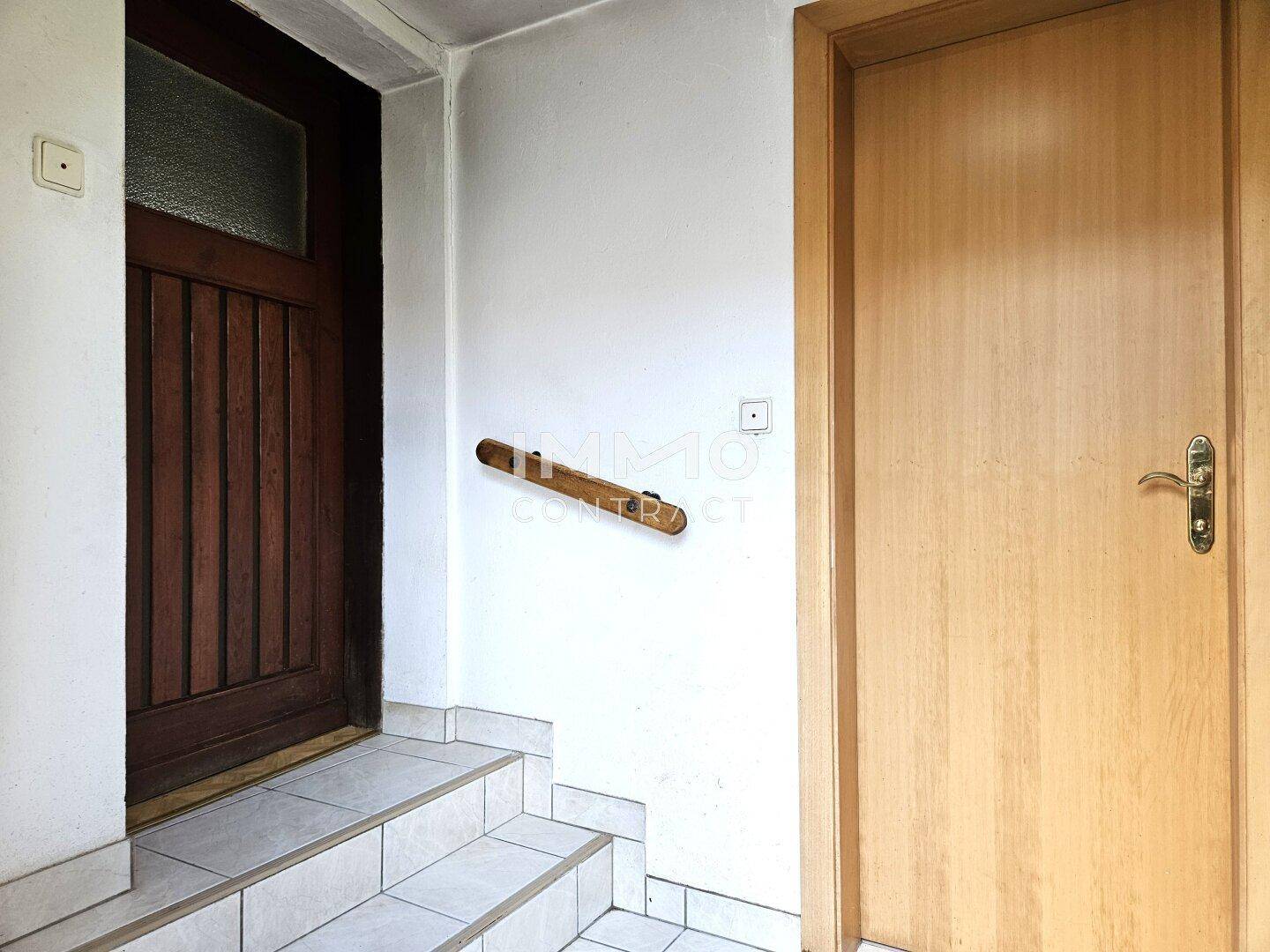Eingangsbereich mit gefliestem Boden, Holztür und Handlauf an der Wand.