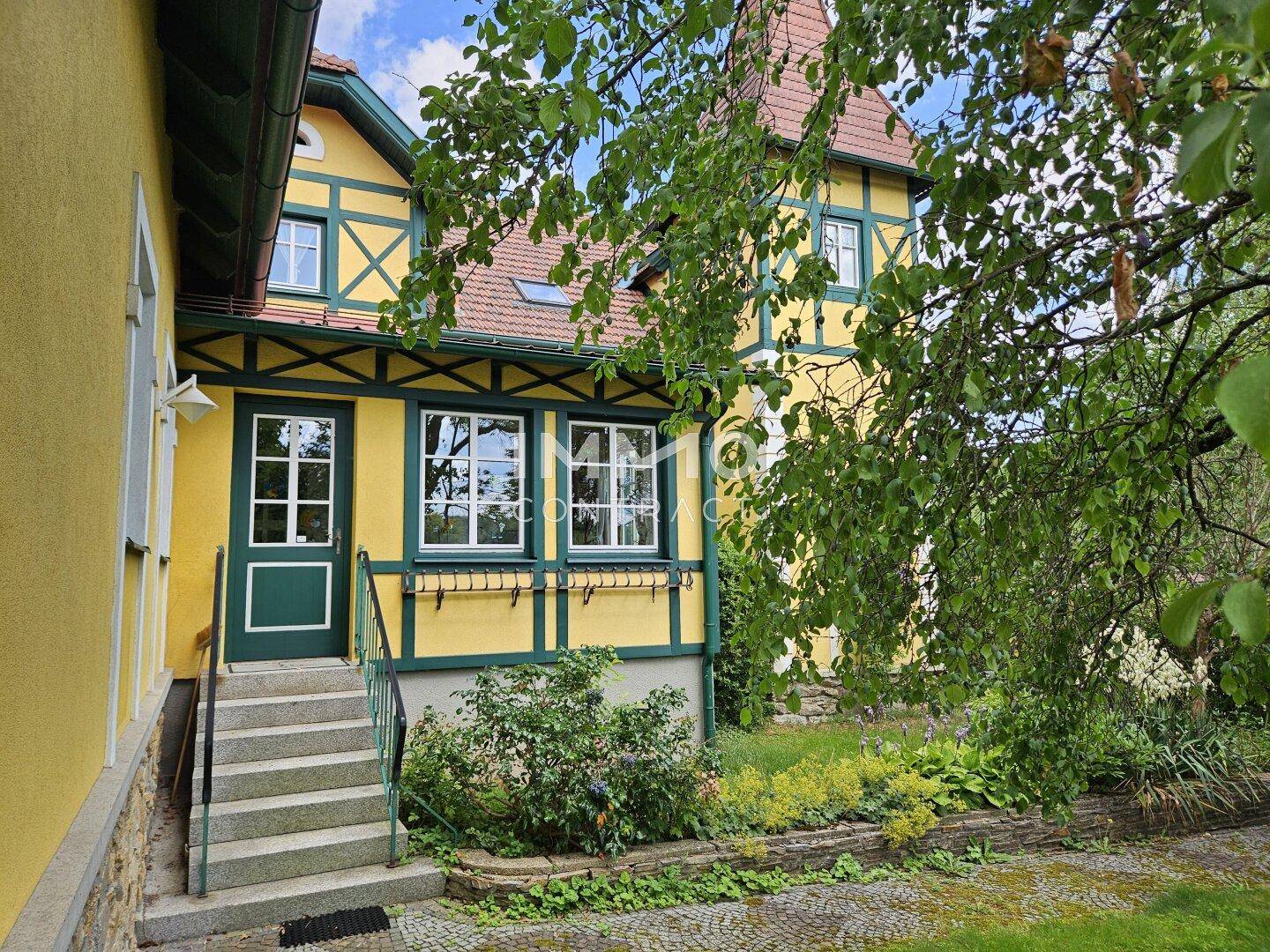 Eingangsbereich des gelben Hauses mit grünen Akzenten, Fenstern und einer kleinen Treppe zum Garten.