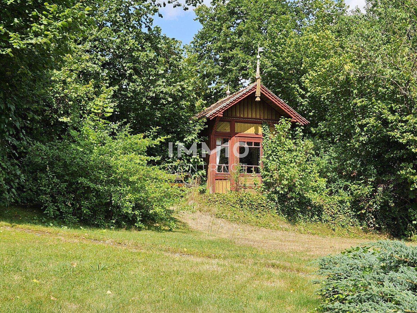 Rustikales Holzhaus oder Pavillon in einem grünen Garten, umgeben von Bäumen und Sträuchern.