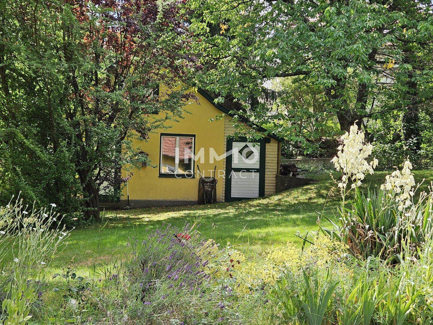 Kleines gelbes Gartenhaus umgeben von dichtem Grün und blühenden Pflanzen in einem weitläufigen Garten.