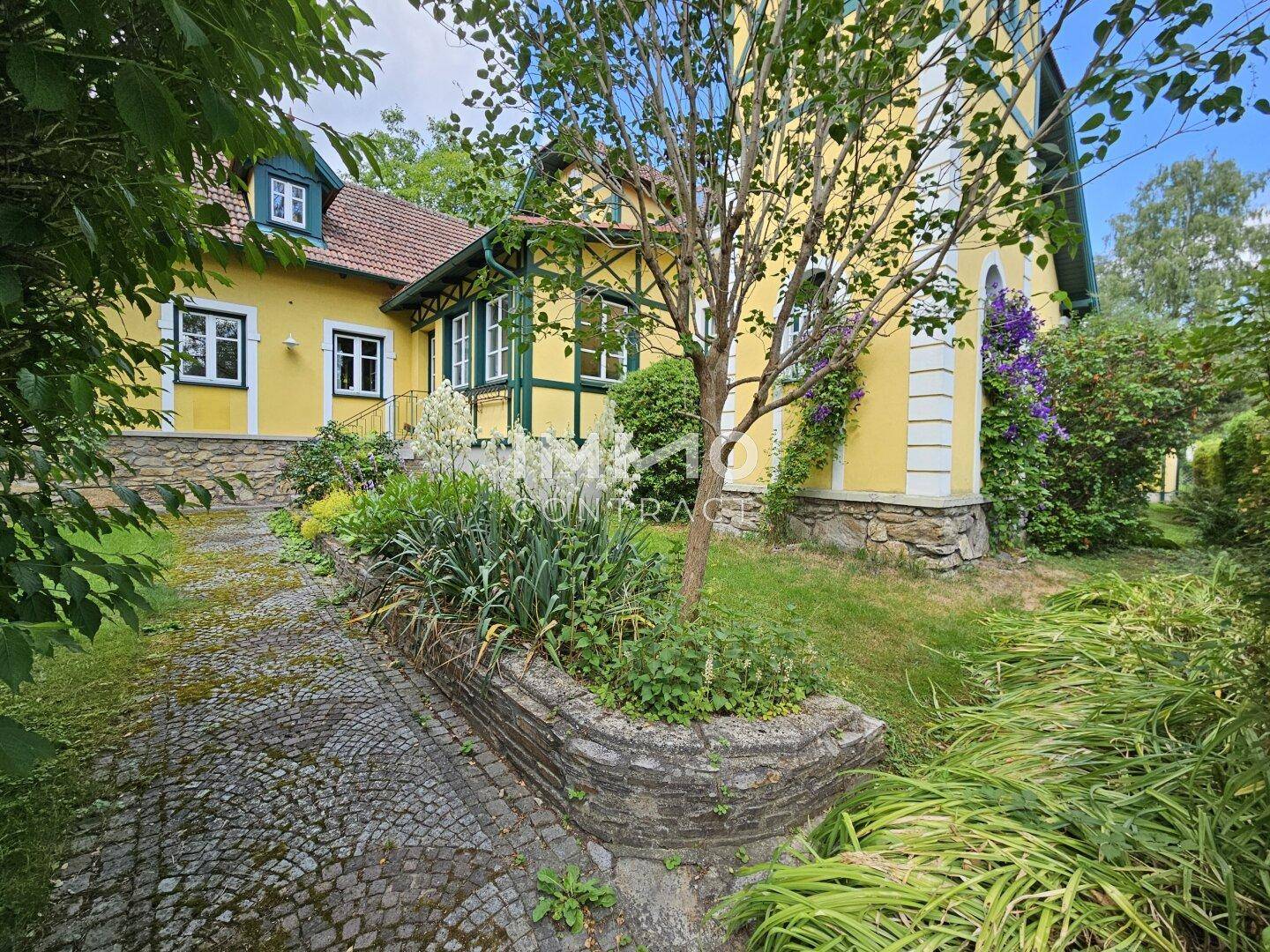 Gartenbereich des Hauses mit gepflastertem Weg, Blumenbeeten und üppiger Vegetation unter blauem Himmel.