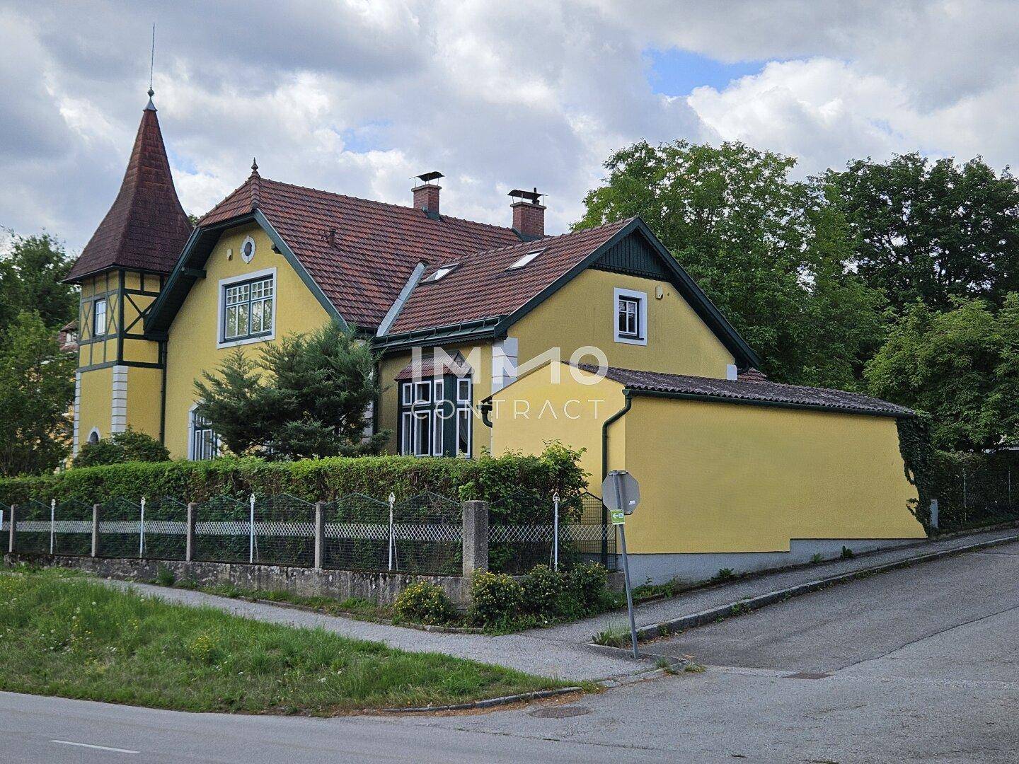Gelbes Haus mit markantem Turm und rotem Ziegeldach, umgeben von Grünflächen an einer Straßenecke.