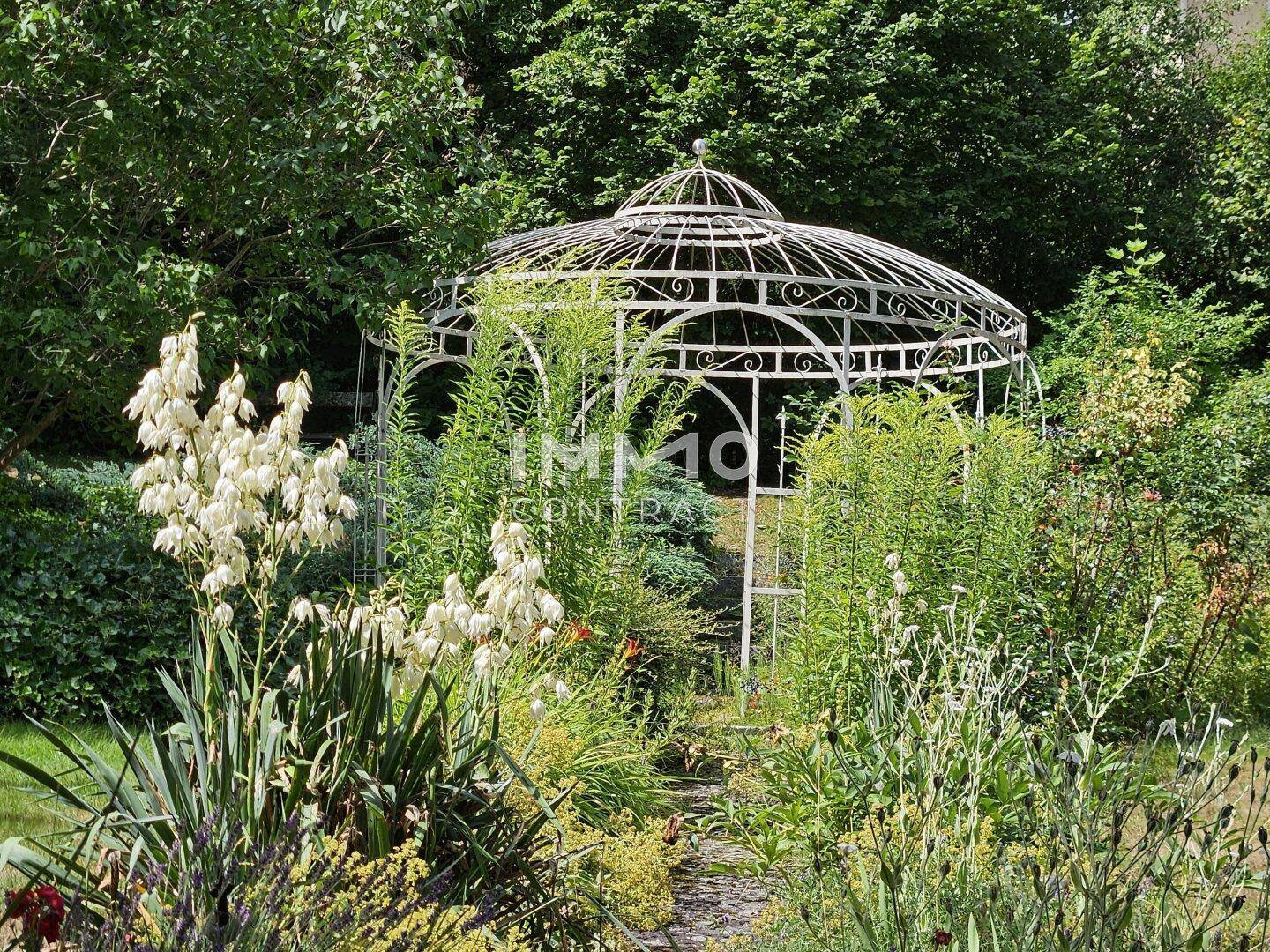 Ein eleganter weißer Metallpavillon steht inmitten eines blühenden Gartens mit hohen Pflanzen und Blumen.