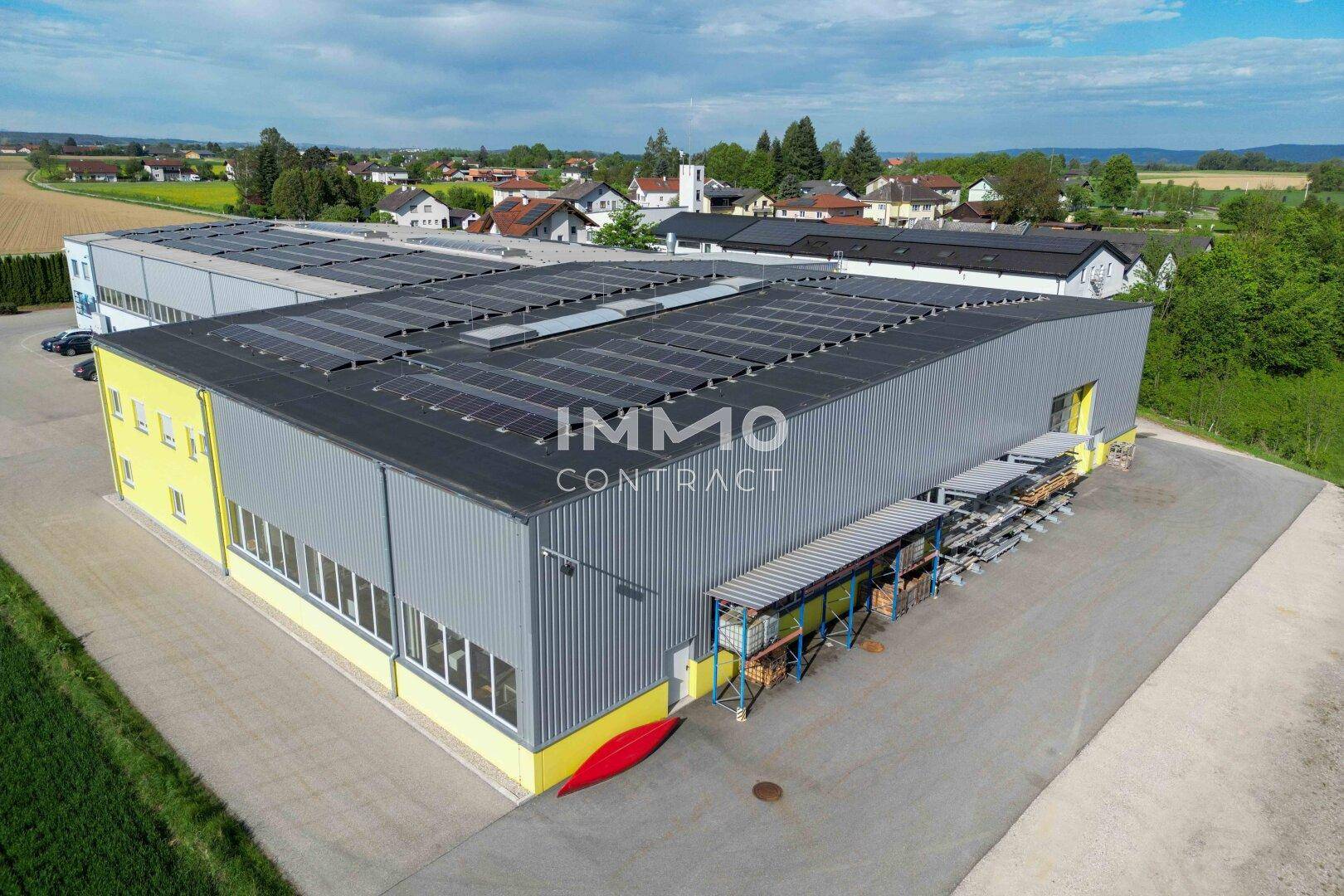 Luftaufnahme einer großen Industriehalle mit Solarmodulen, gelegen am Rande einer ländlichen Siedlung mit Wohnhäusern.