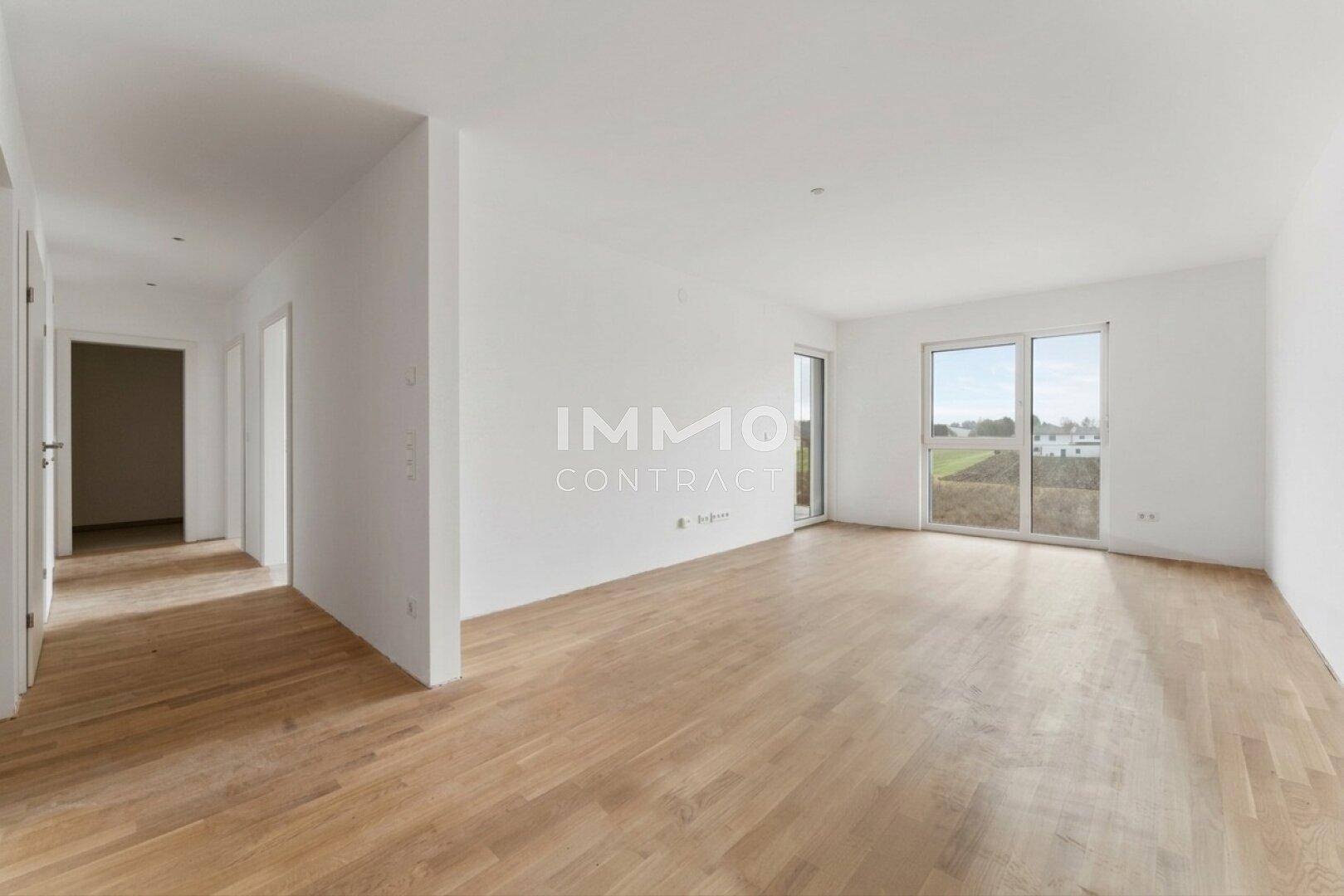 Geräumiges, leeres Wohnzimmer mit großen Fenstern, Holzboden und Blick ins Grüne.