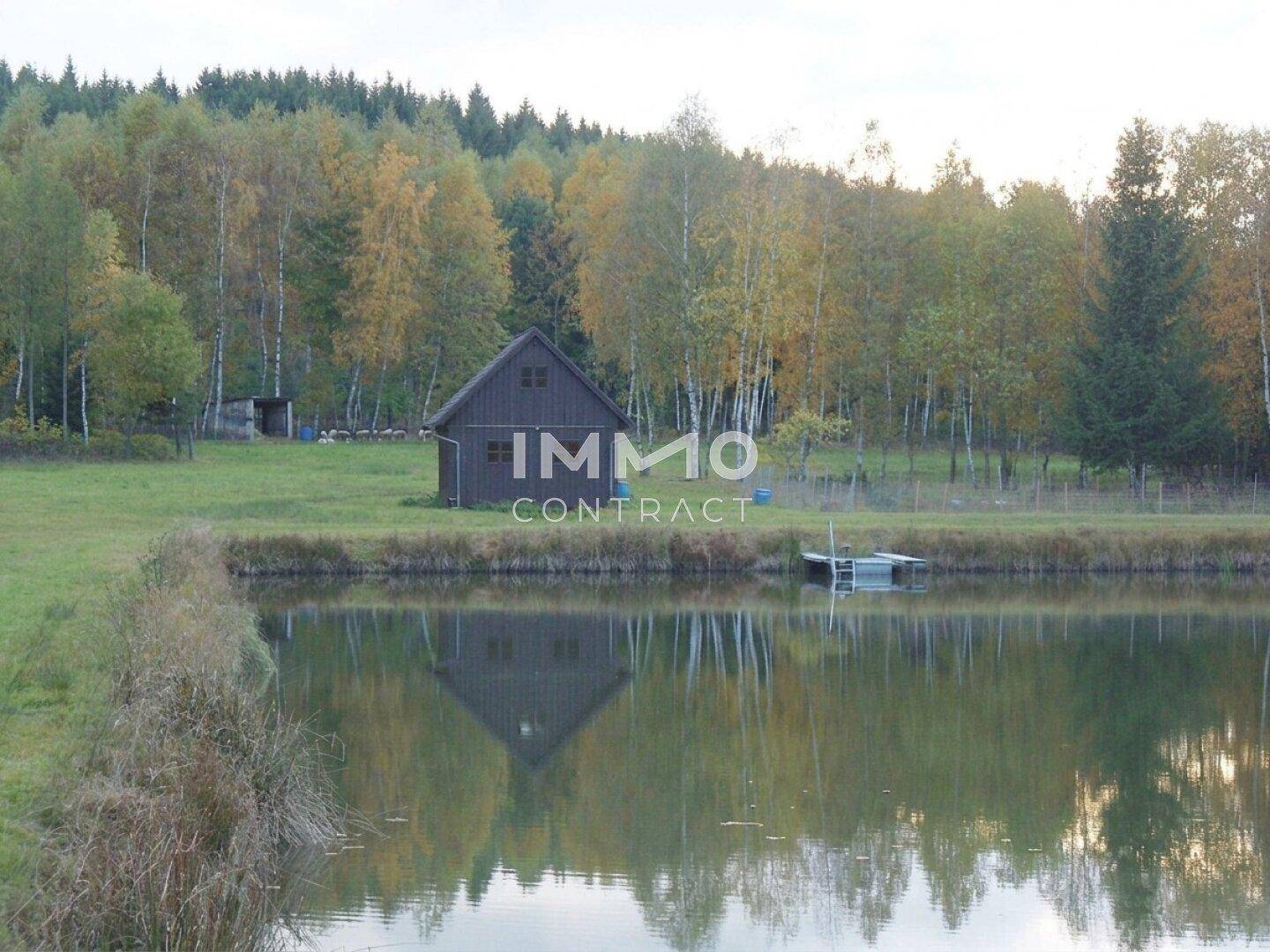 Teich mit Spiegelung eines Holzhauses und herbstlicher Bäume am gegenüberliegenden Ufer.