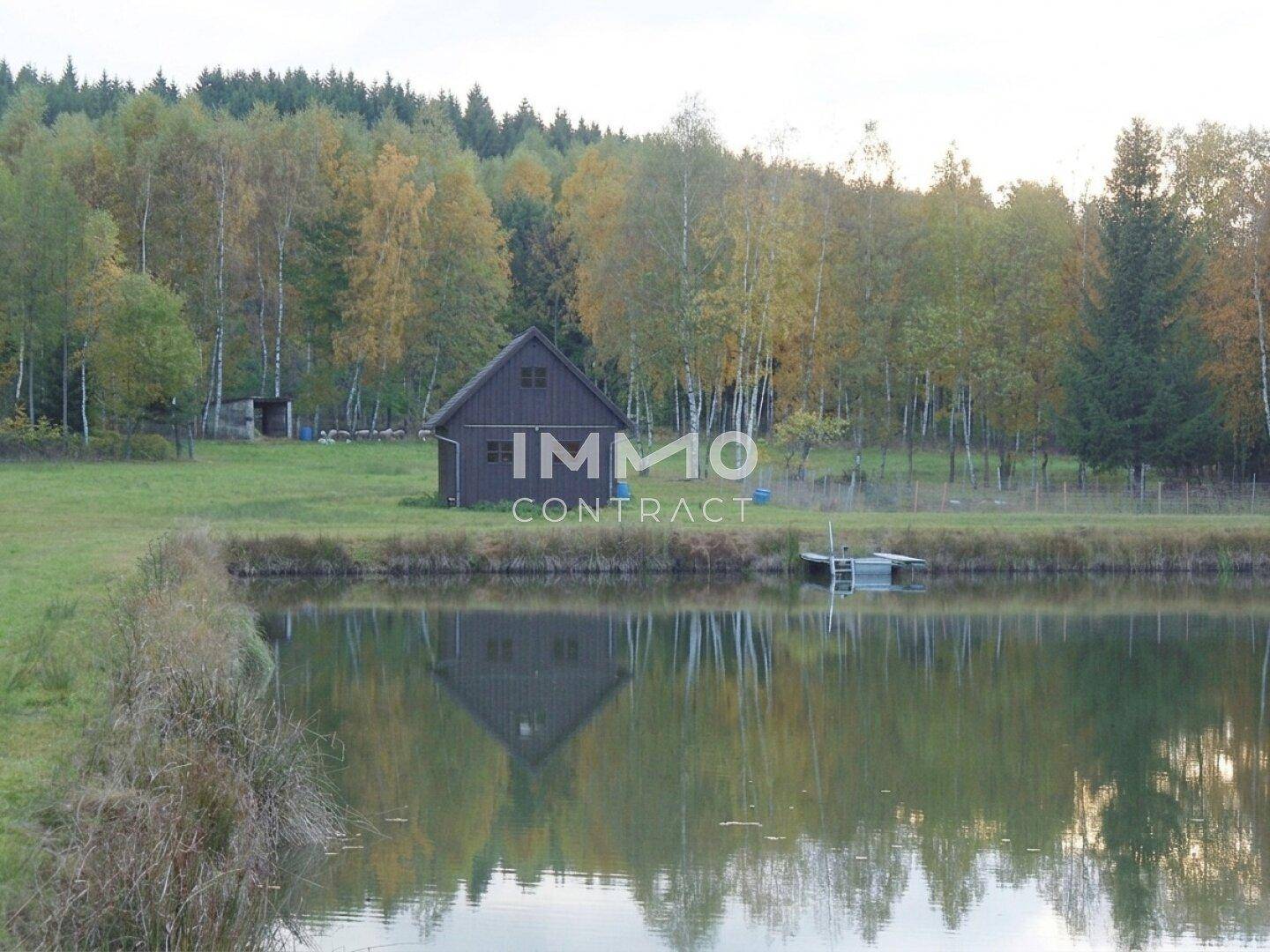 Rustikales Holzhaus am Ufer eines Teiches, umgeben von Bäumen in herbstlicher Farbenpracht.