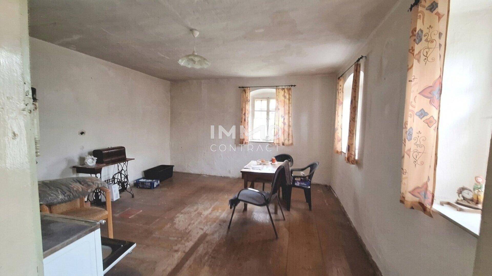 Einfacher Essbereich mit Holzdielenboden, zwei Fenstern und einem Tisch mit Stühlen in ursprünglichem Zustand.