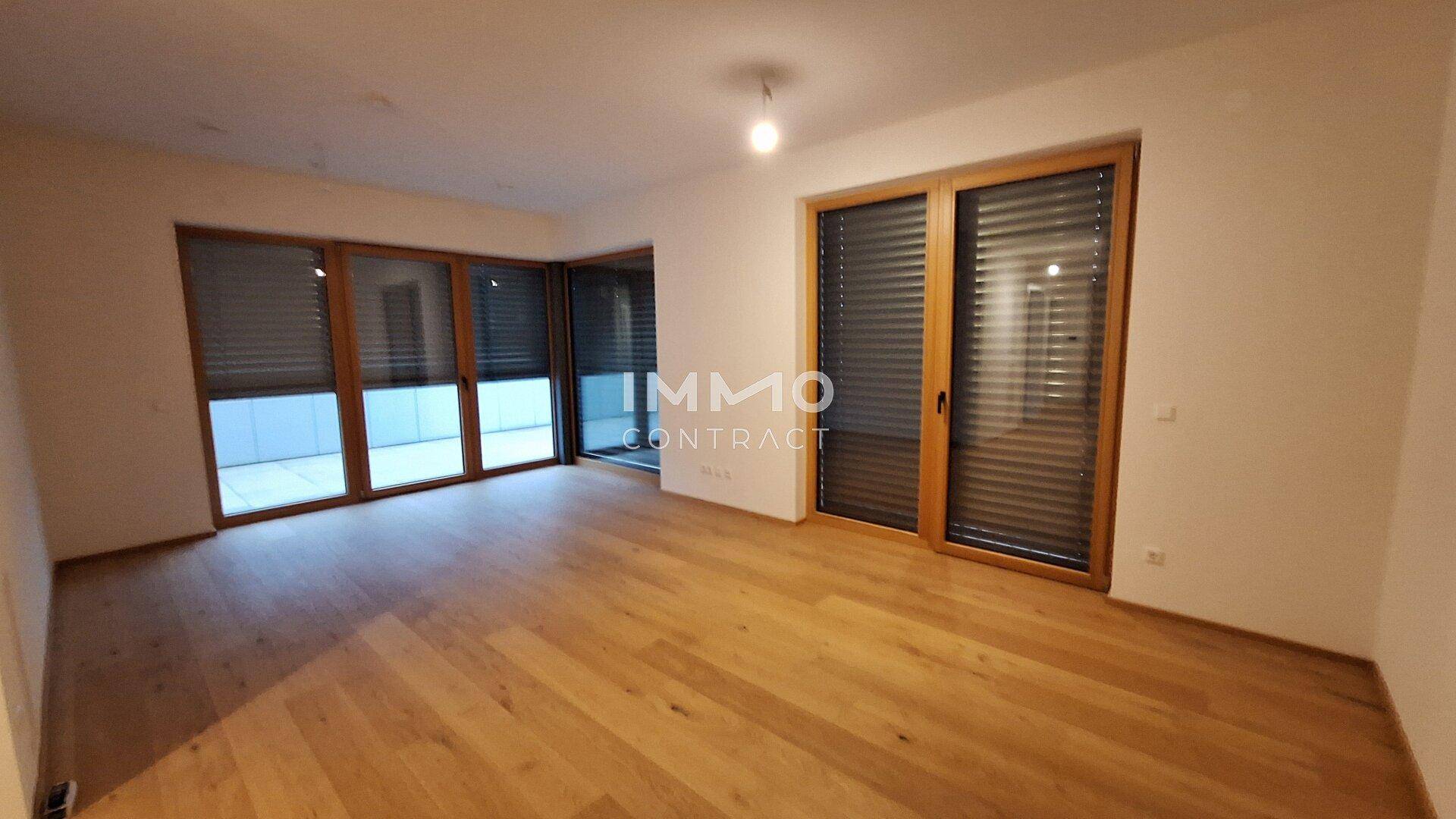 Heller, leerer Wohnraum mit hochwertigem Holzparkett und großen Fenstern mit Jalousien.