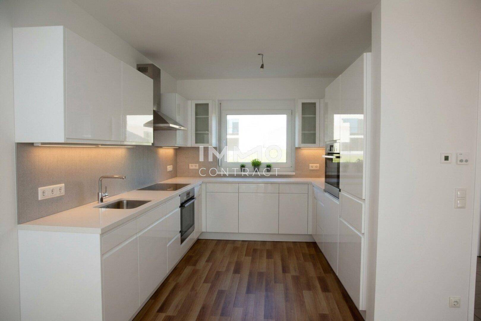 Moderne weiße Einbauküche mit glänzenden Fronten und einem Fenster über der Spüle.