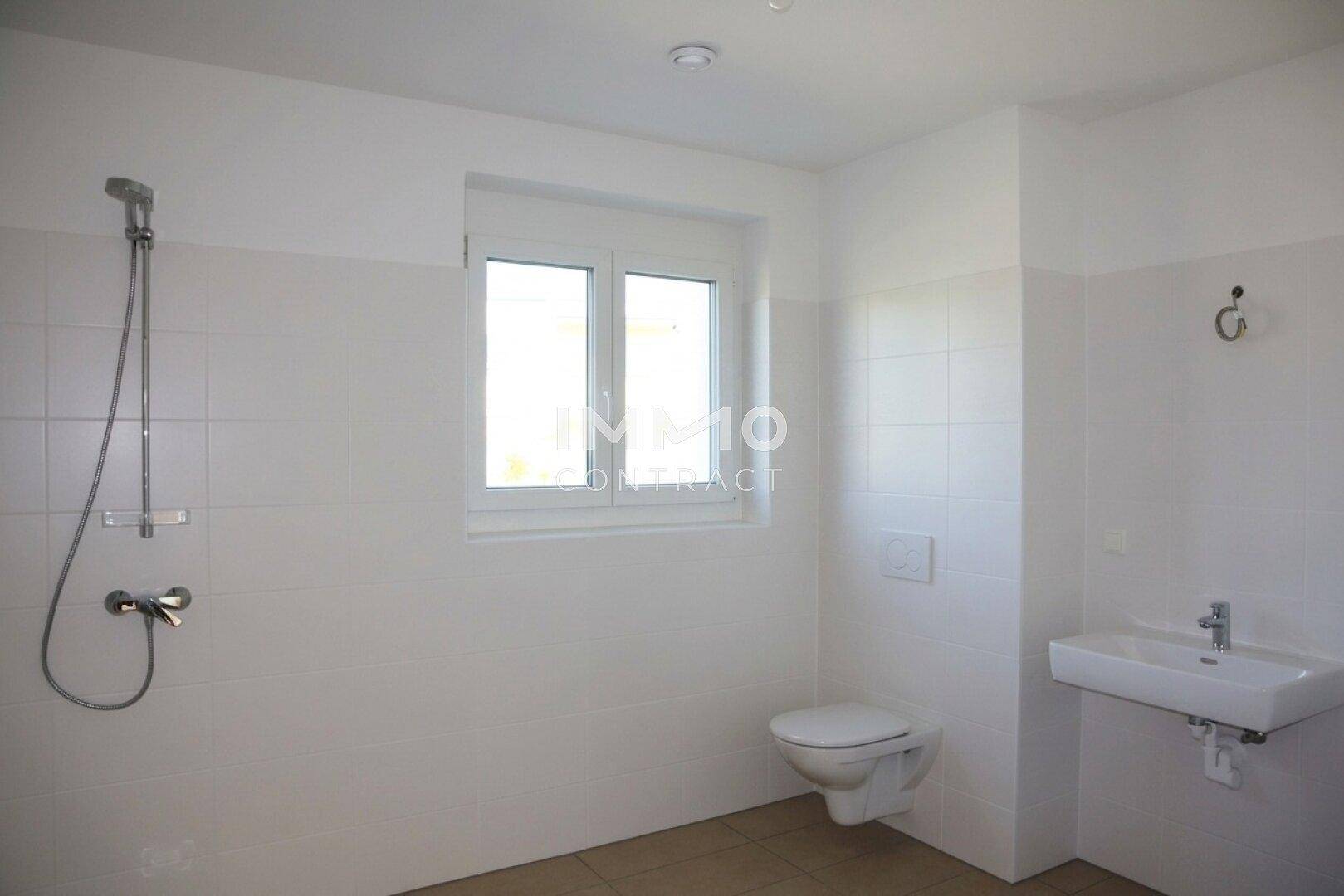 Modernes Badezimmer mit weißen Fliesen, Dusche, WC, Waschbecken und Fenster.
