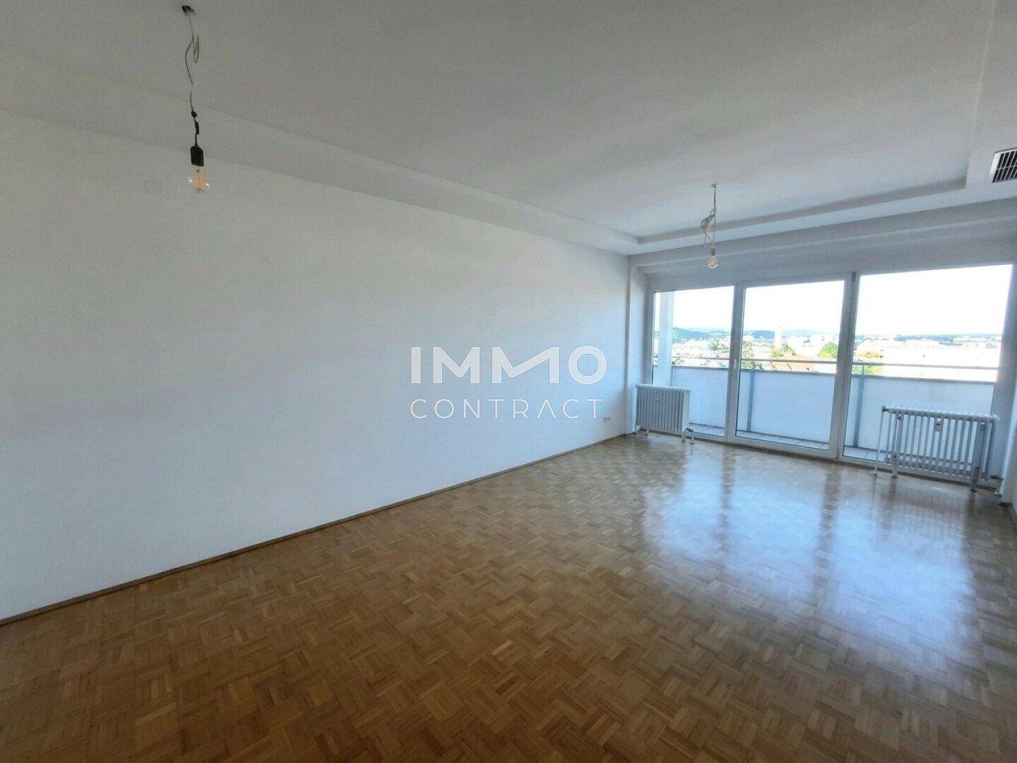 Helles, leeres Wohnzimmer mit Fischgrätparkett und großen Fenstern, die viel Tageslicht hereinlassen.