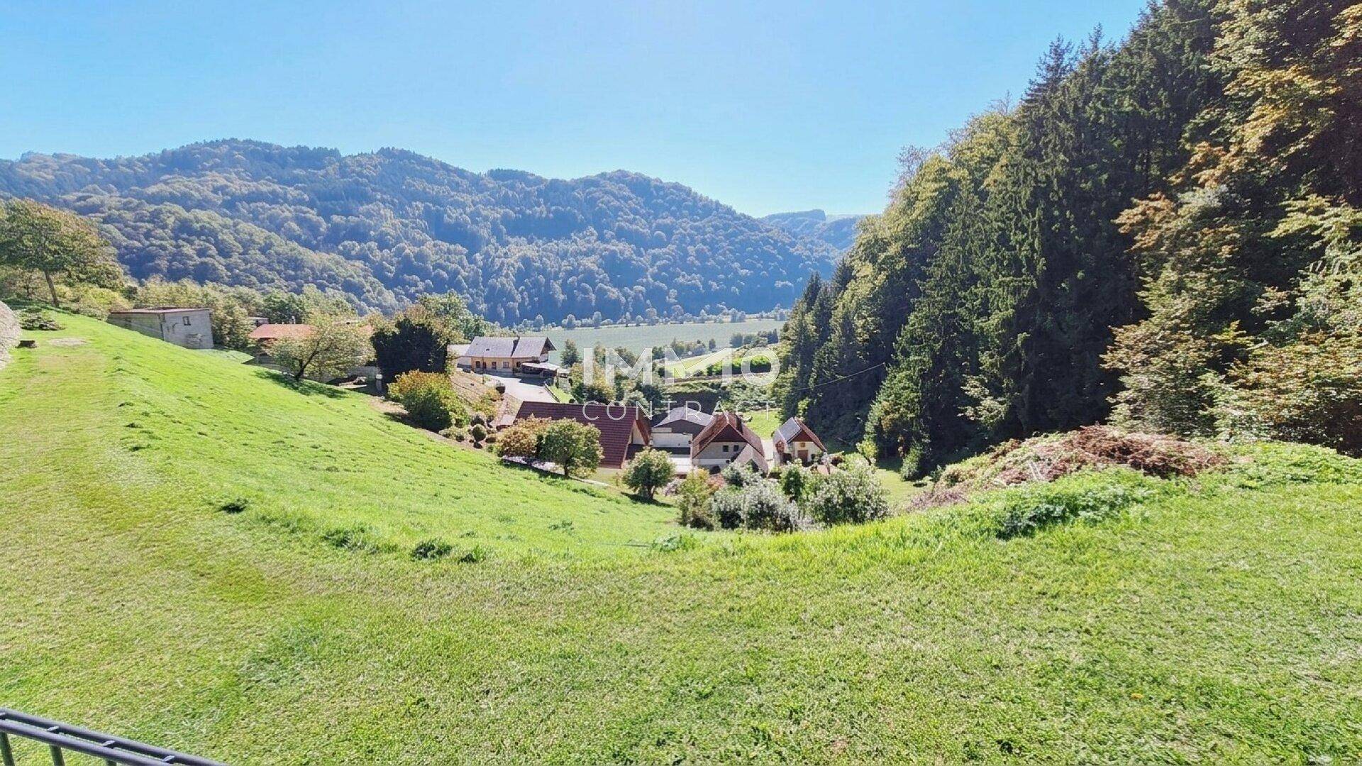 Weitläufiger Blick über eine grüne Hügellandschaft mit vereinzelten Häusern und einem See im Hintergrund.
