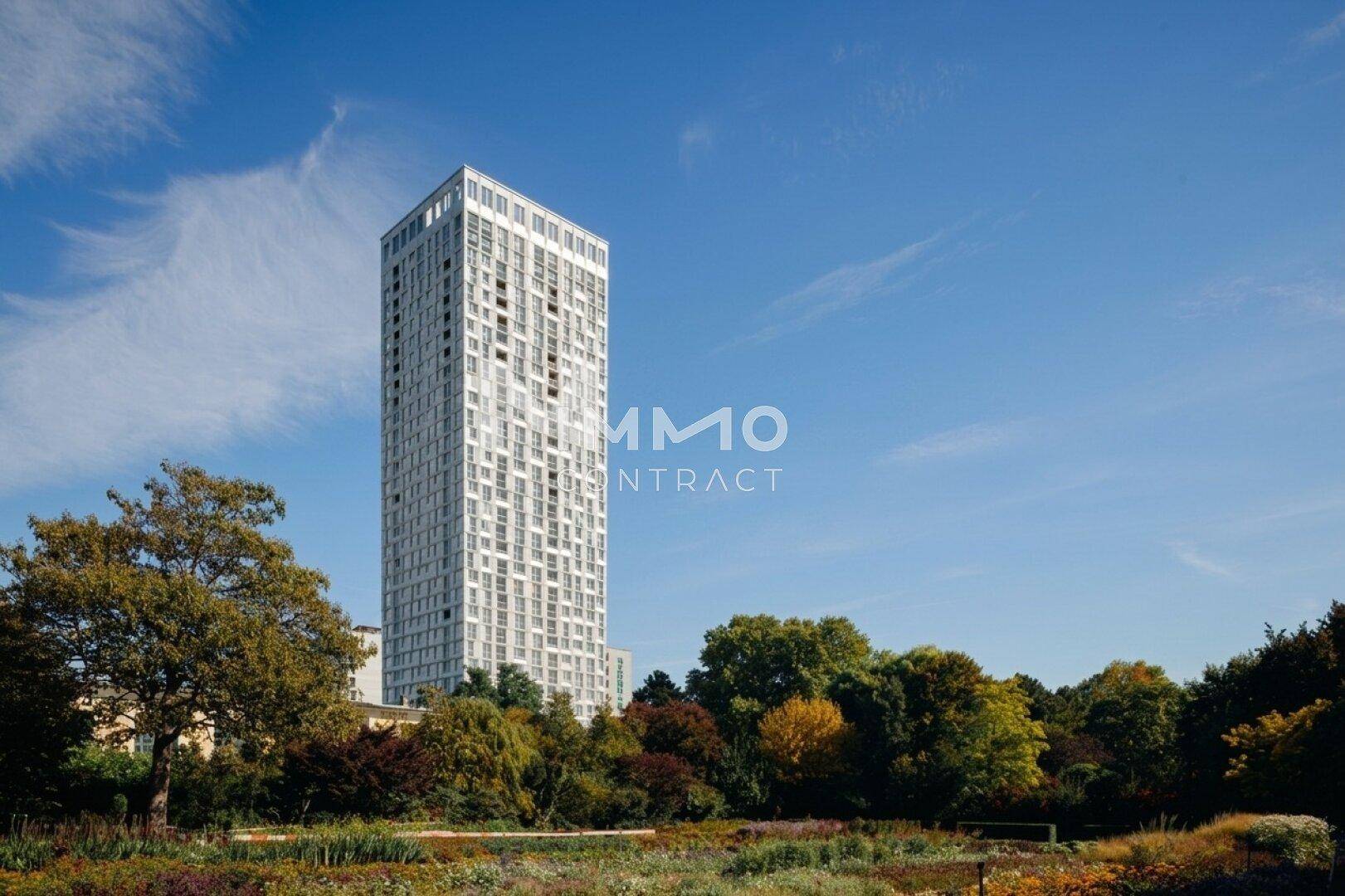Moderner Wolkenkratzer umgeben von üppiger Natur und einem weitläufigen Park unter blauem Himmel.