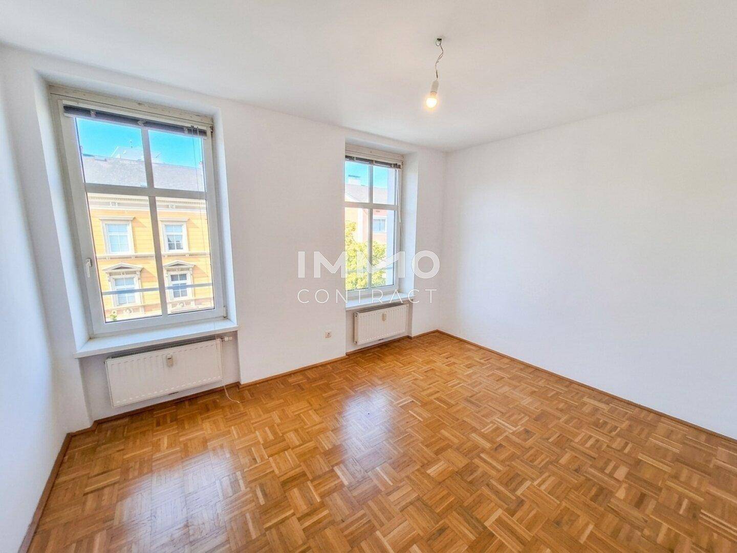 Helles, leeres Zimmer mit Fischgrätparkett und zwei Fenstern, ideal als Schlafzimmer.