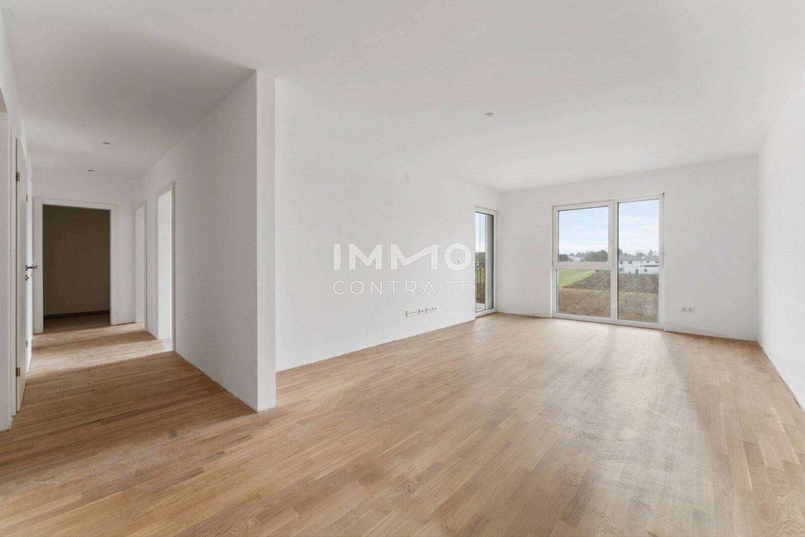 Heller, unmöblierter Wohnraum mit Holzboden und großen Fenstern mit Blick ins Grüne.