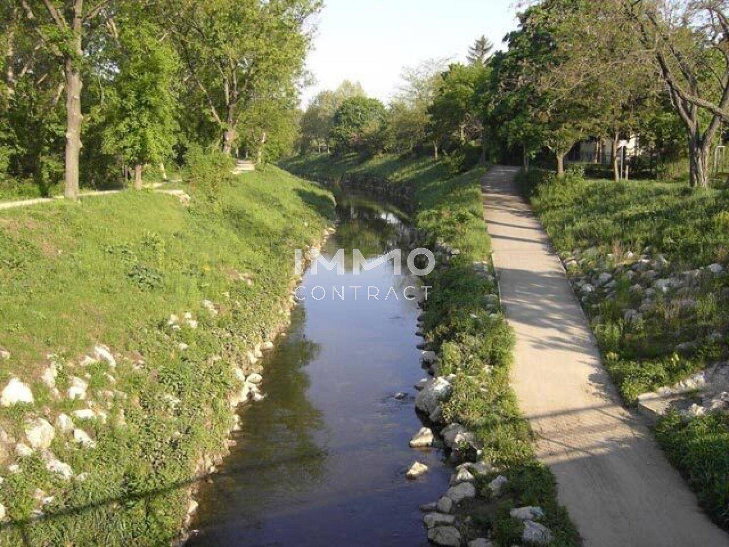 Malerischer Flusslauf mit befestigtem Uferweg, umgeben von üppigem Grün und Bäumen.