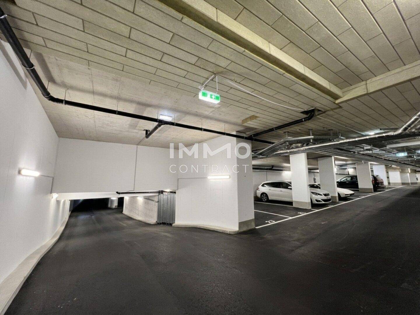 Heller Eingangsbereich einer Tiefgarage mit Rampe und geparkten Fahrzeugen.