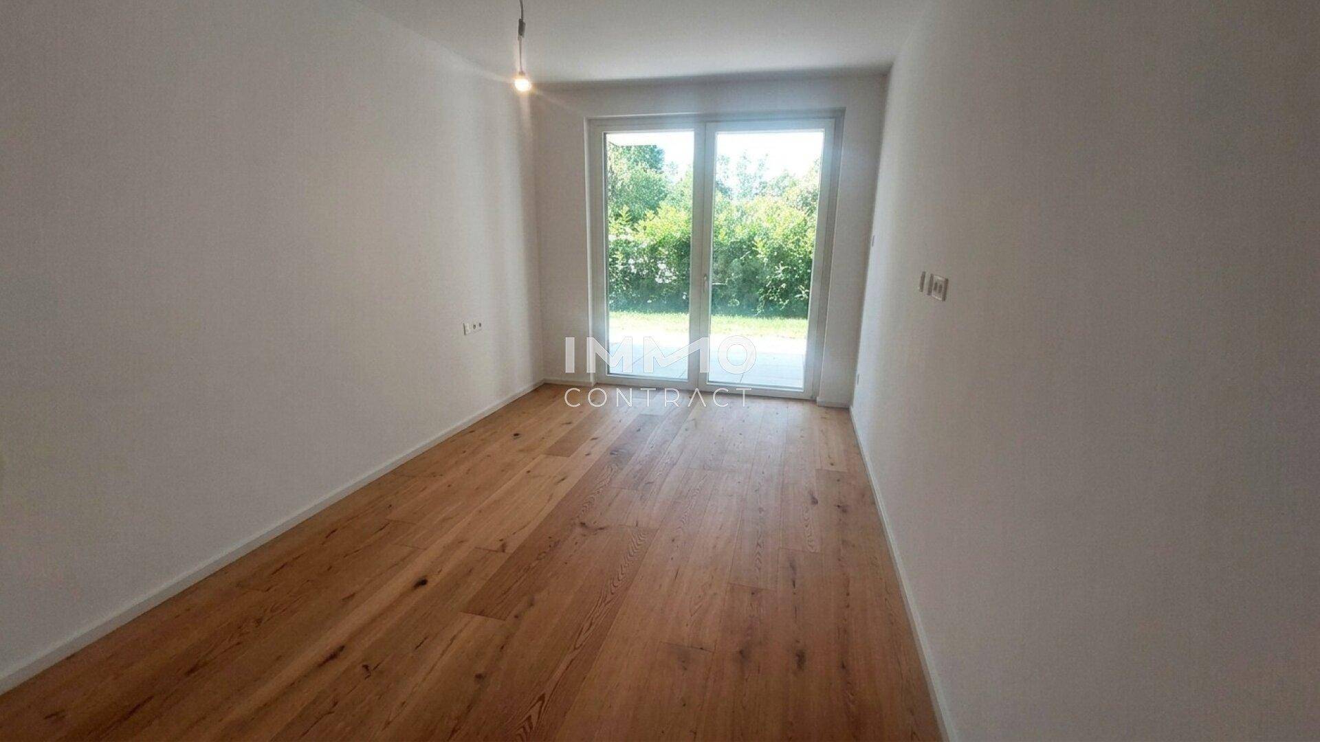 Helles Zimmer mit Holzboden und großer Glastür, die zu einem grünen Außenbereich führt.