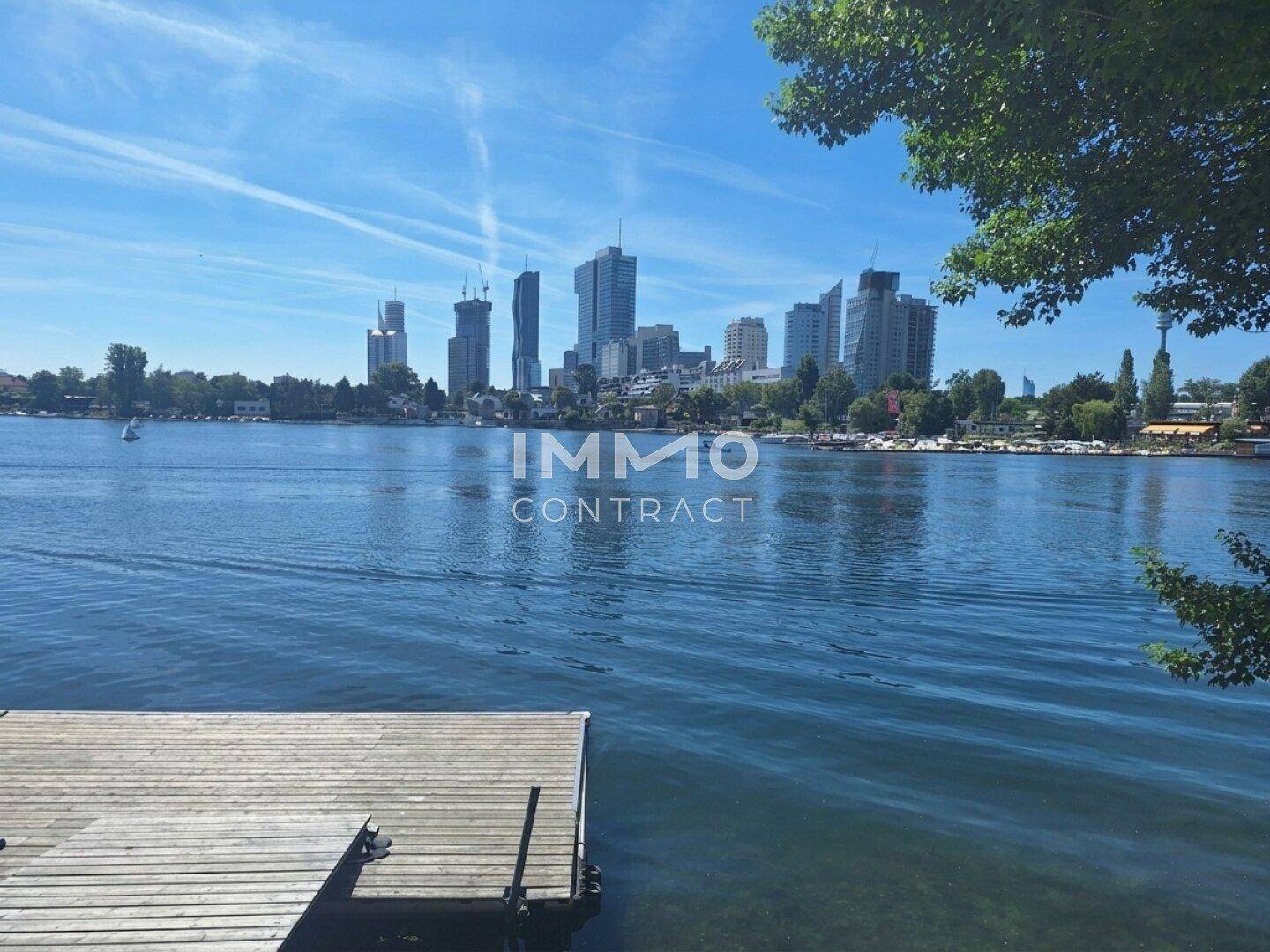 Panoramablick auf den See und die Stadtkulisse mit einem Holzsteg im Vordergrund an einem sonnigen Tag.