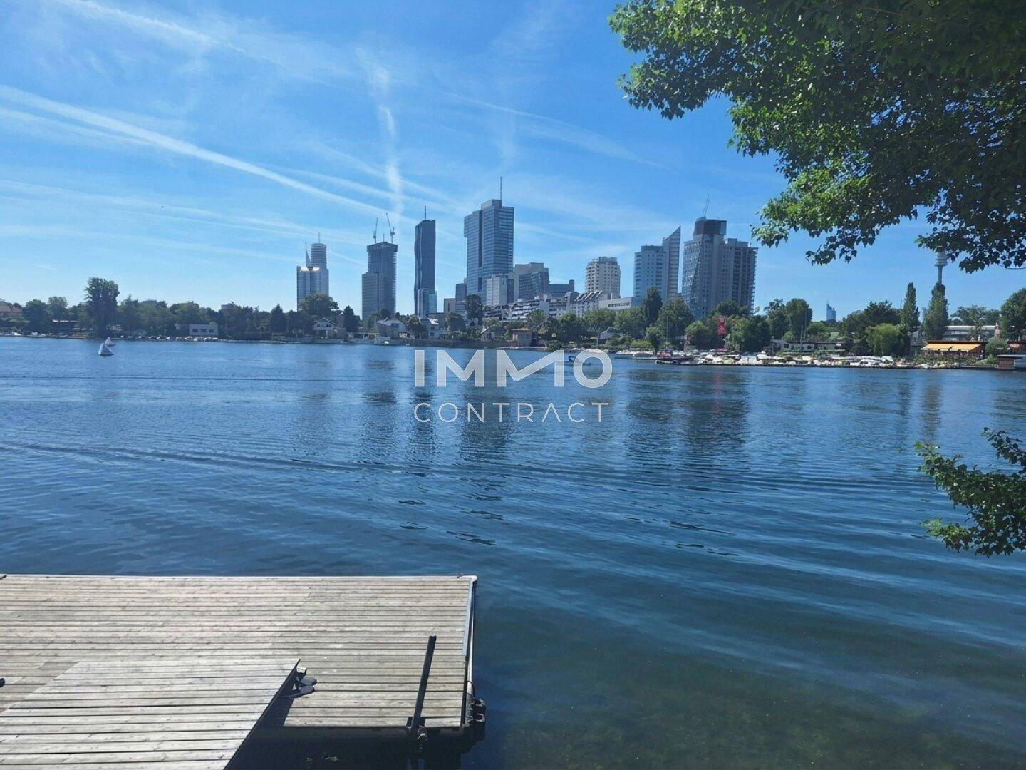 Weitläufiger Seeblick mit Stadtpanorama und einem Holzsteg im Vordergrund unter blauem Himmel.