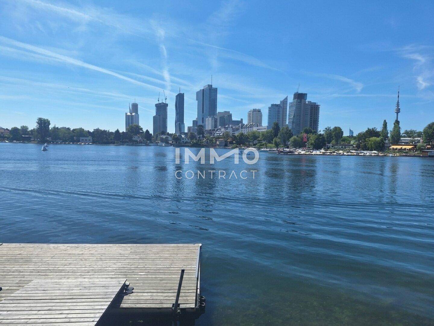 Weitläufiger Blick über den See mit einem Holzsteg im Vordergrund und der Stadtkulisse am Horizont.