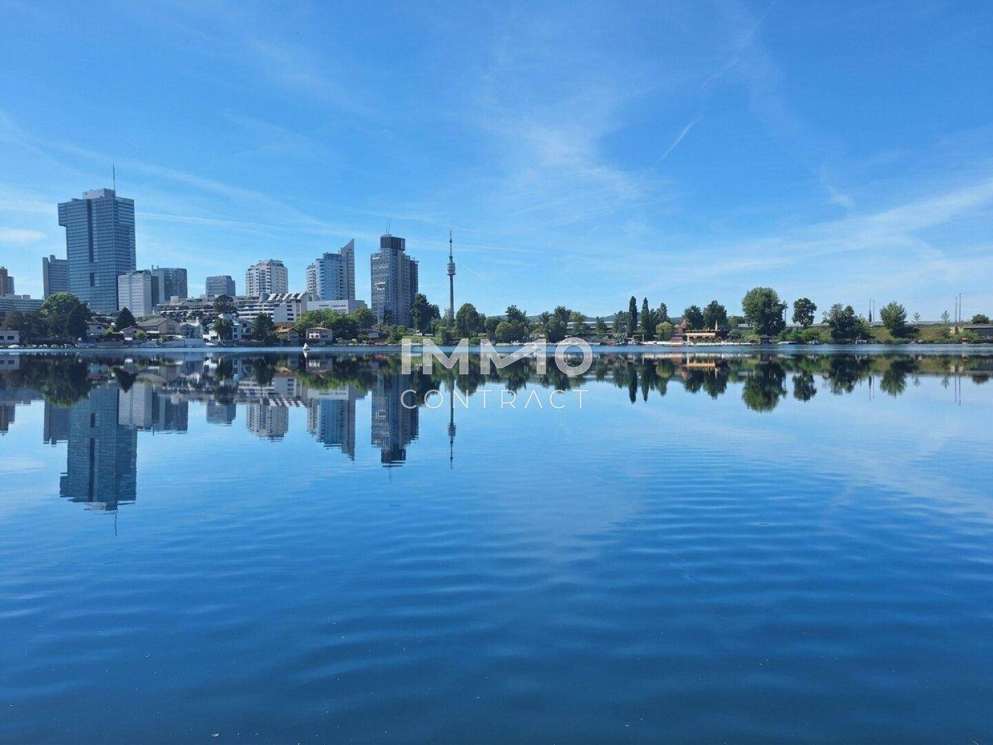 Panoramablick über den See mit der Skyline einer modernen Stadt im Hintergrund unter blauem Himmel.