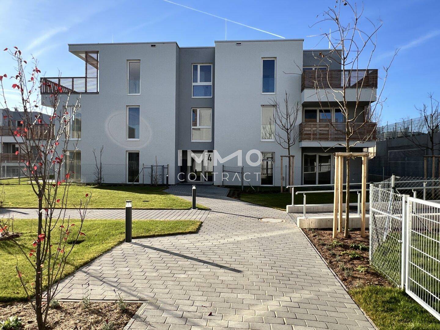 Vorderansicht eines modernen Mehrfamilienhauses mit Zugangsweg und Grünflächen im Sonnenschein.