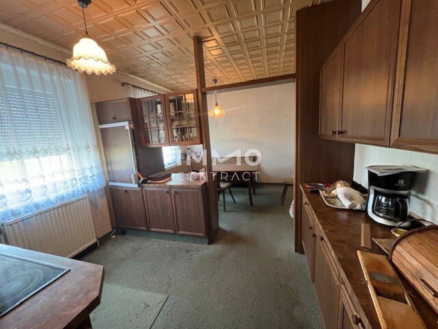 Ältere Küche mit Holzschränken, Teppichboden und einem kleinen Essbereich im Hintergrund.