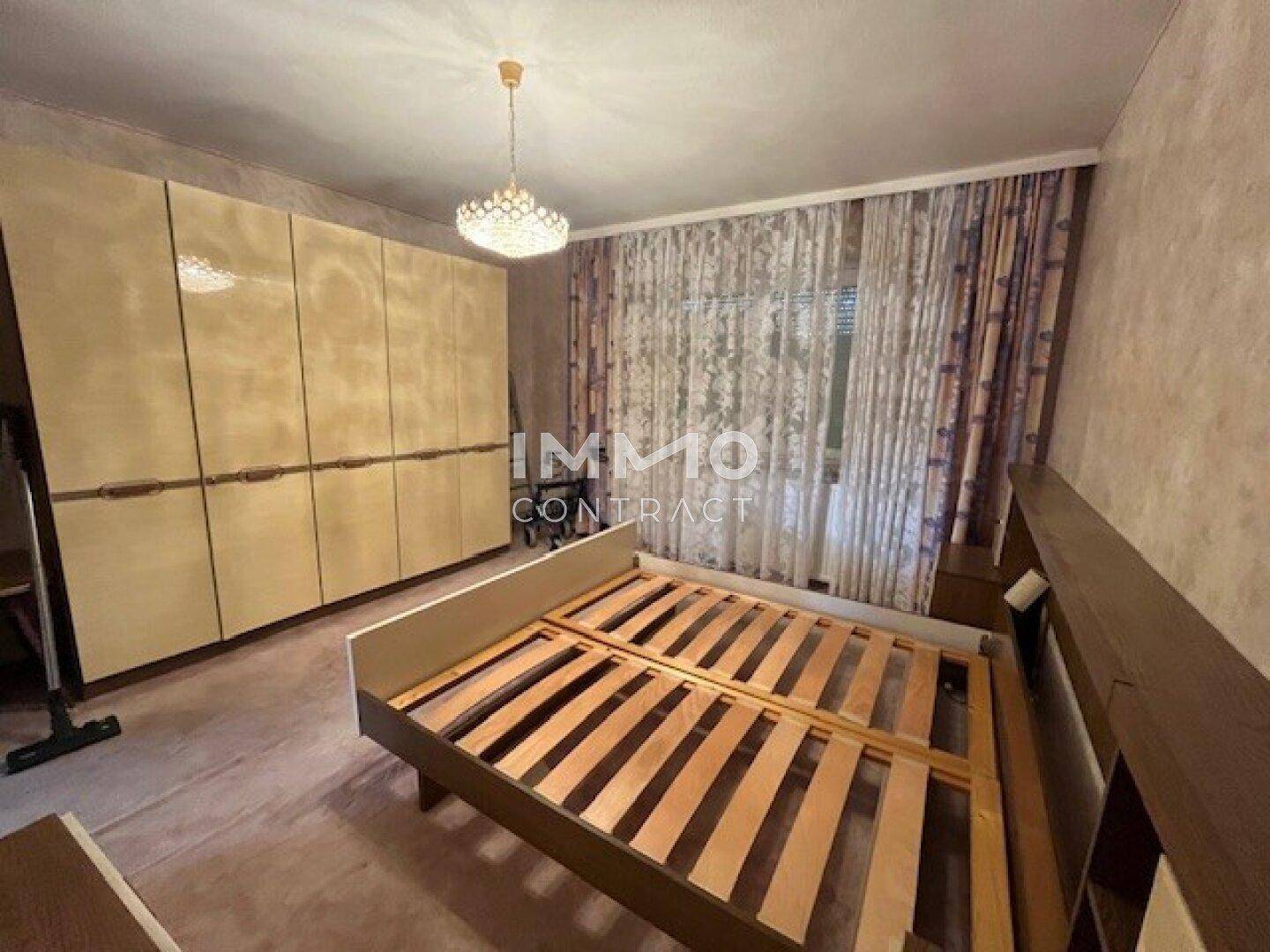 Schlafzimmer mit großem Kleiderschrank, Bettrahmen und gemusterten Vorhängen vor dem Fenster.