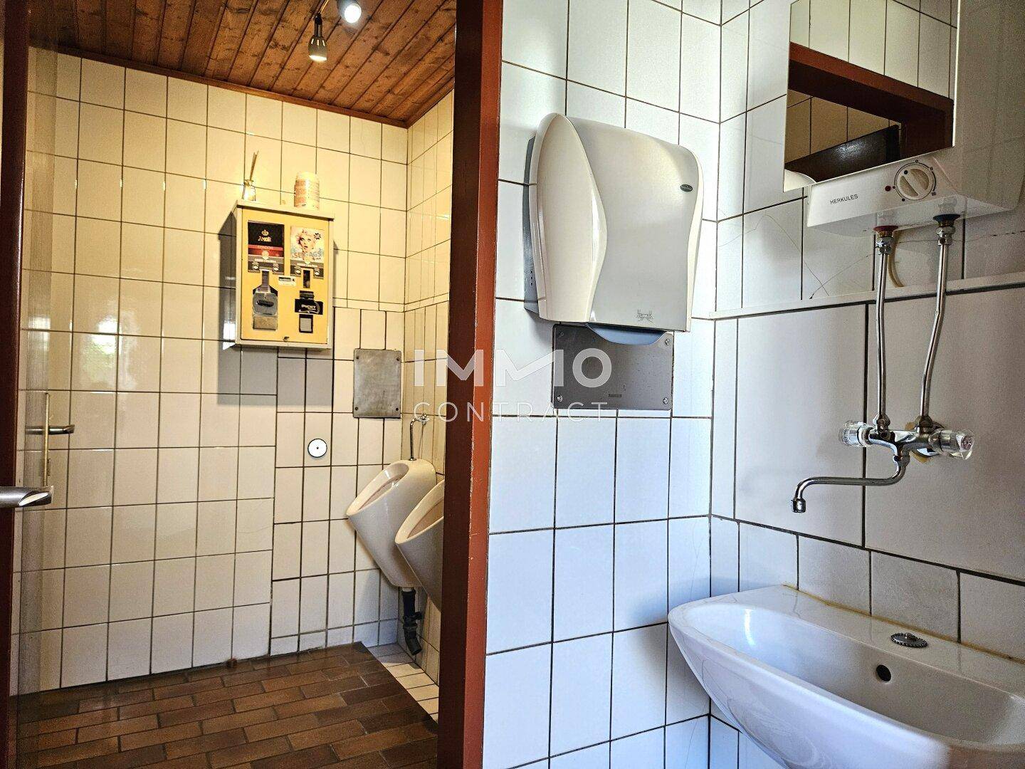 Herrentoilette mit Urinalen, Waschbecken und weißen Fliesen, ausgestattet mit Handtuchspender.