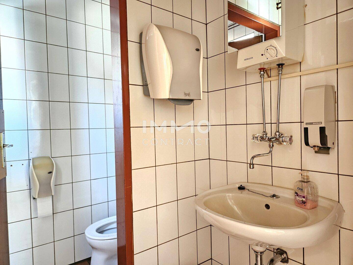Waschbereich der Toilette mit Waschbecken, Spiegel und weißen Fliesen, sauber und funktional.