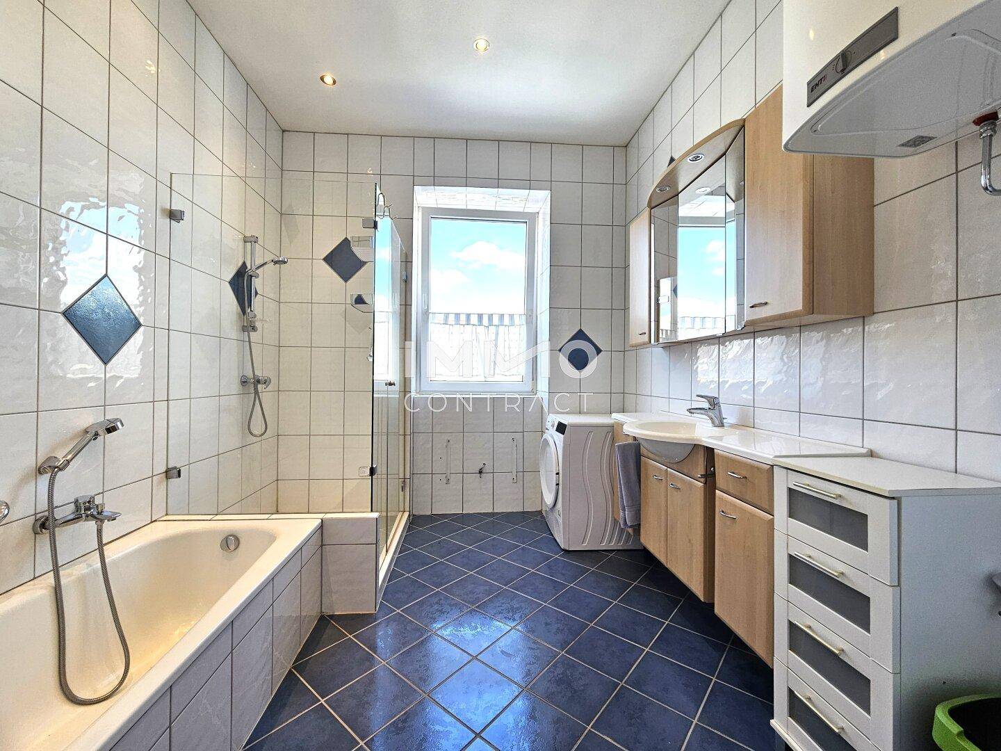 Badezimmer mit Badewanne, Dusche, Waschmaschine und Fenster, komplett gefliest in Weiß und Blau.