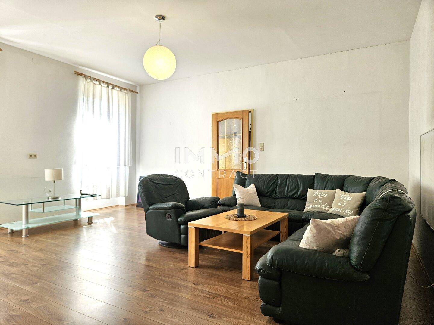 Geräumiges Wohnzimmer mit dunkler Ledercouch, Holzboden und großen Fenstern, hell und einladend.