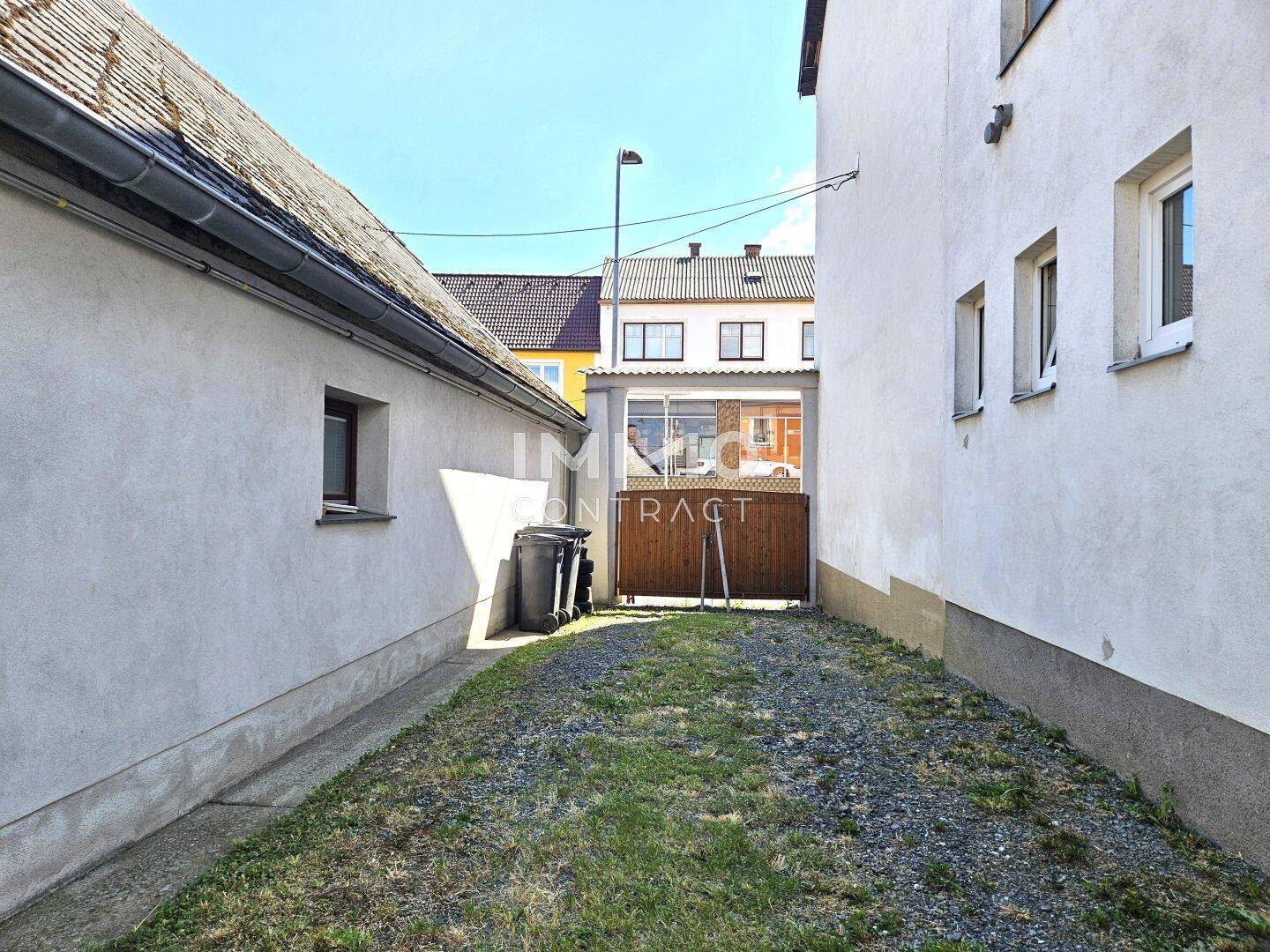 Schmaler Kiesweg zwischen zwei Gebäuden, der zu einem Holztor und weiteren Gebäuden führt.