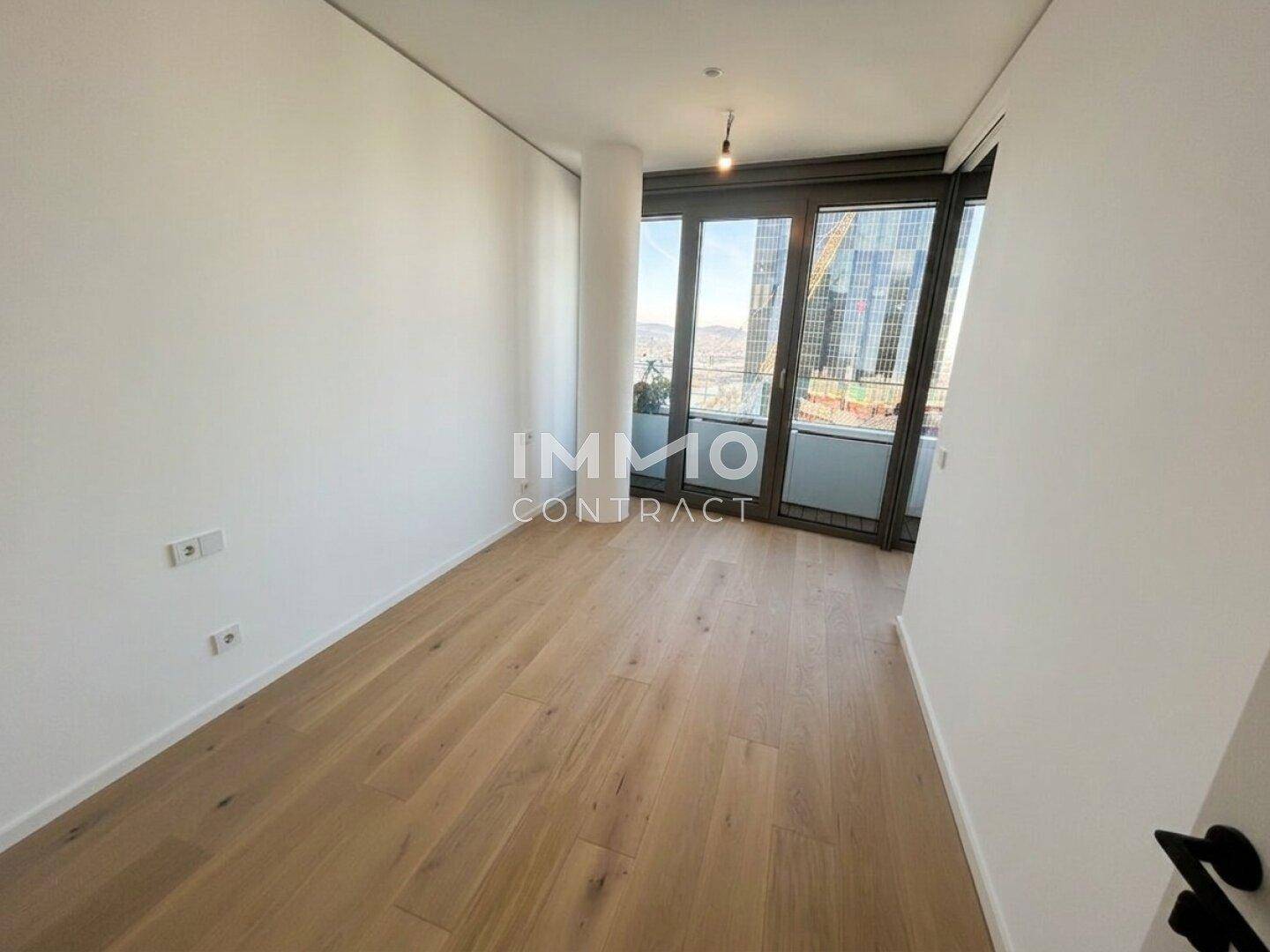 Helles, leeres Zimmer mit Holzboden und großen Fenstern mit Stadtblick.