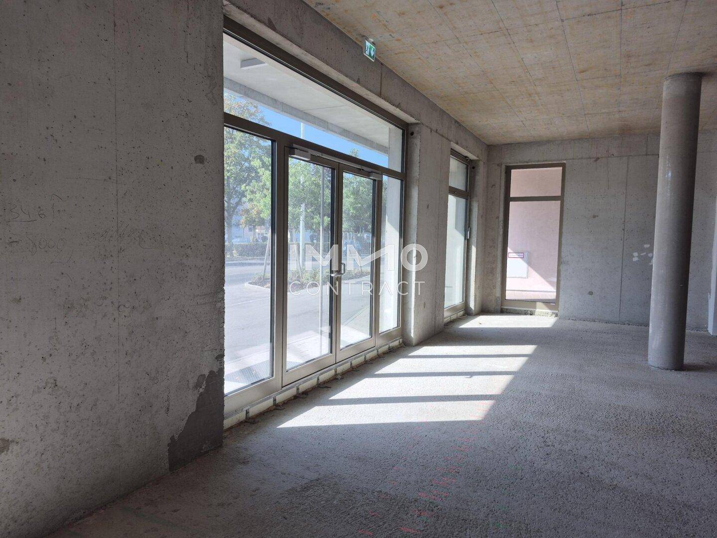 Unfertiger Gewerberaum mit großen Glasfronten, die viel Tageslicht hereinlassen und einen Blick nach draußen bieten.
