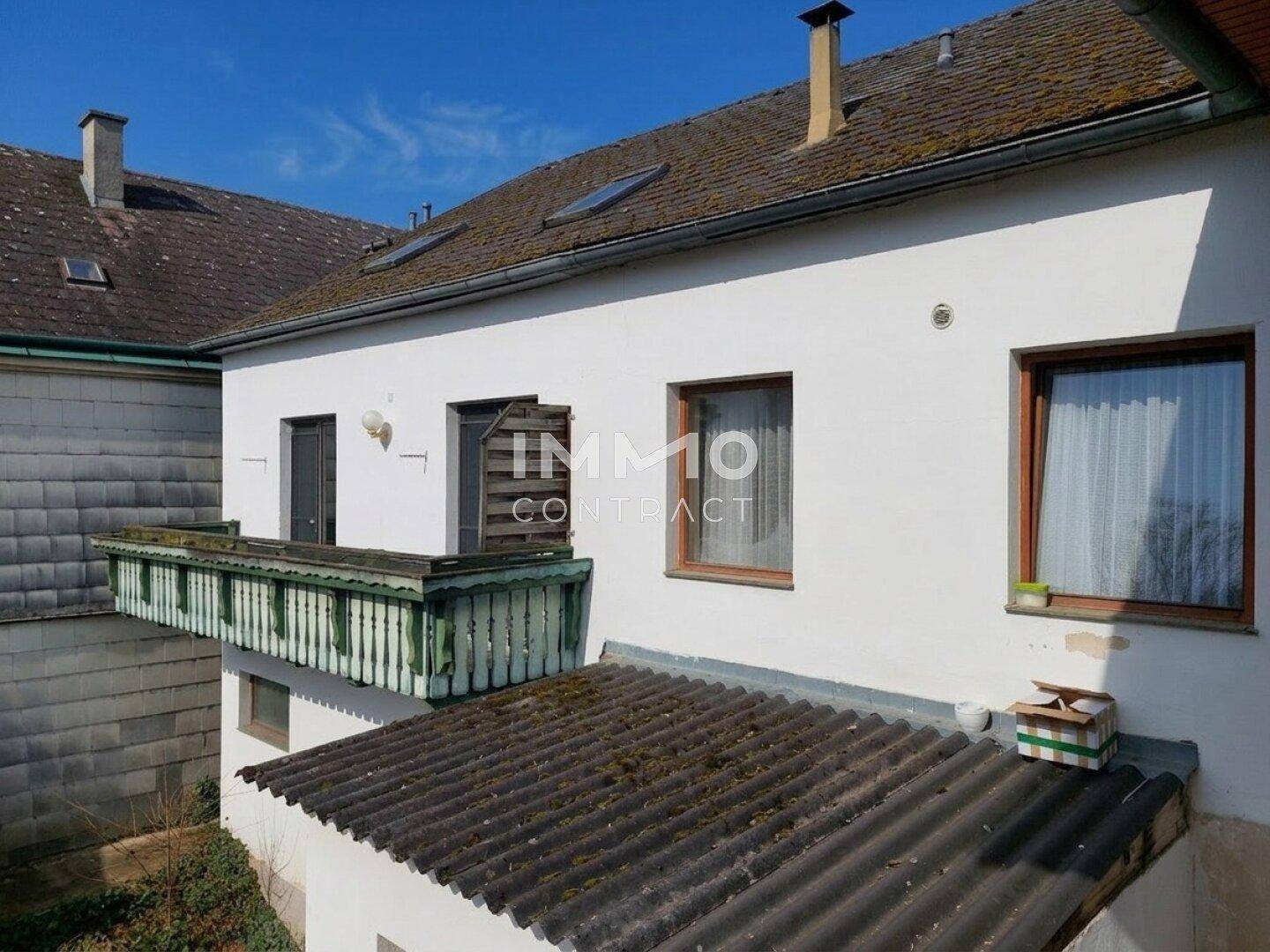 Weißes Gebäude mit traditionellem Holzbalkon und altem Ziegeldach unter blauem Himmel.