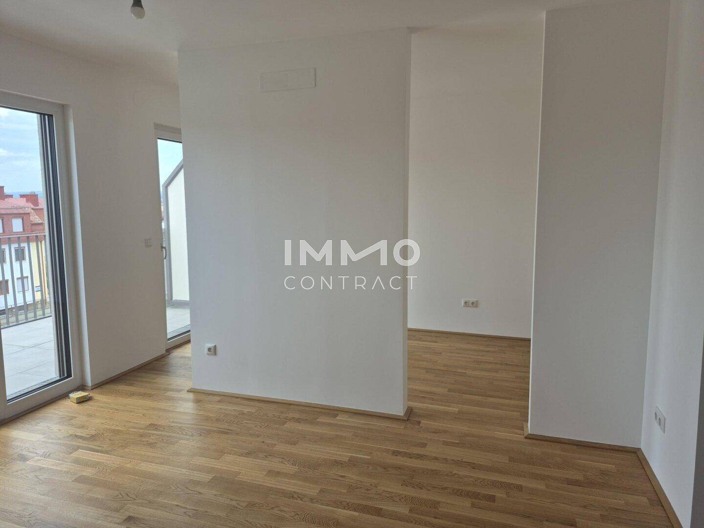 Geräumiger, heller Raum mit Holzboden und großen Glastüren, die zu einem Außenbereich führen.