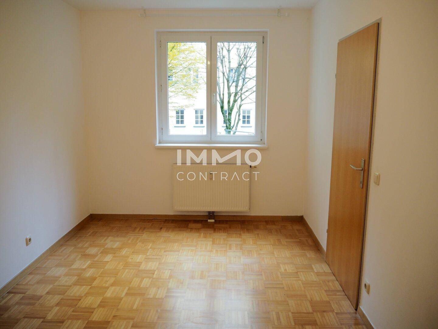 Schlafzimmer mit Parkettboden und großem Fenster, das viel Tageslicht hereinlässt.