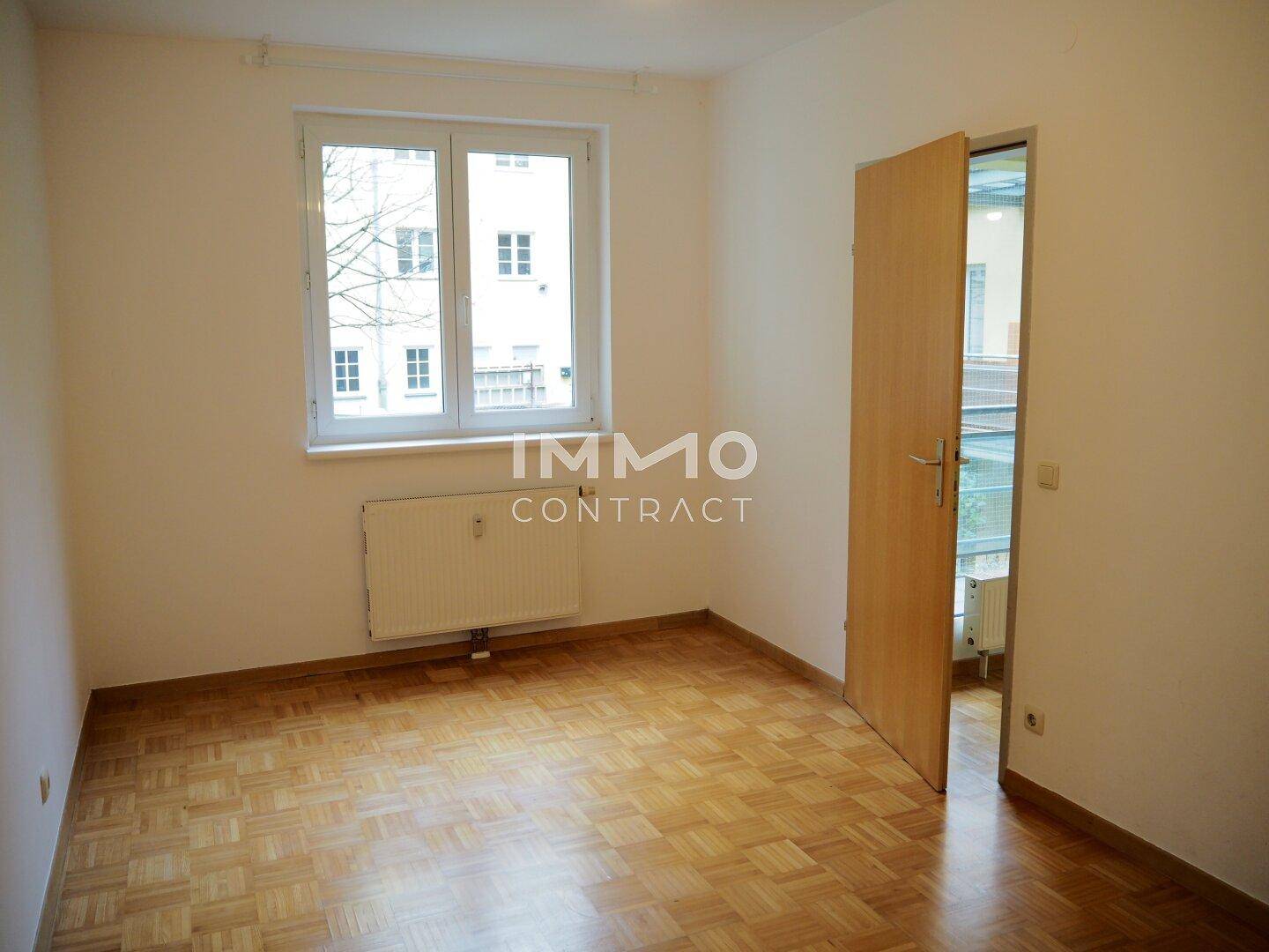 Helles Schlafzimmer mit Parkettboden und einem Fenster mit Blick ins Grüne.