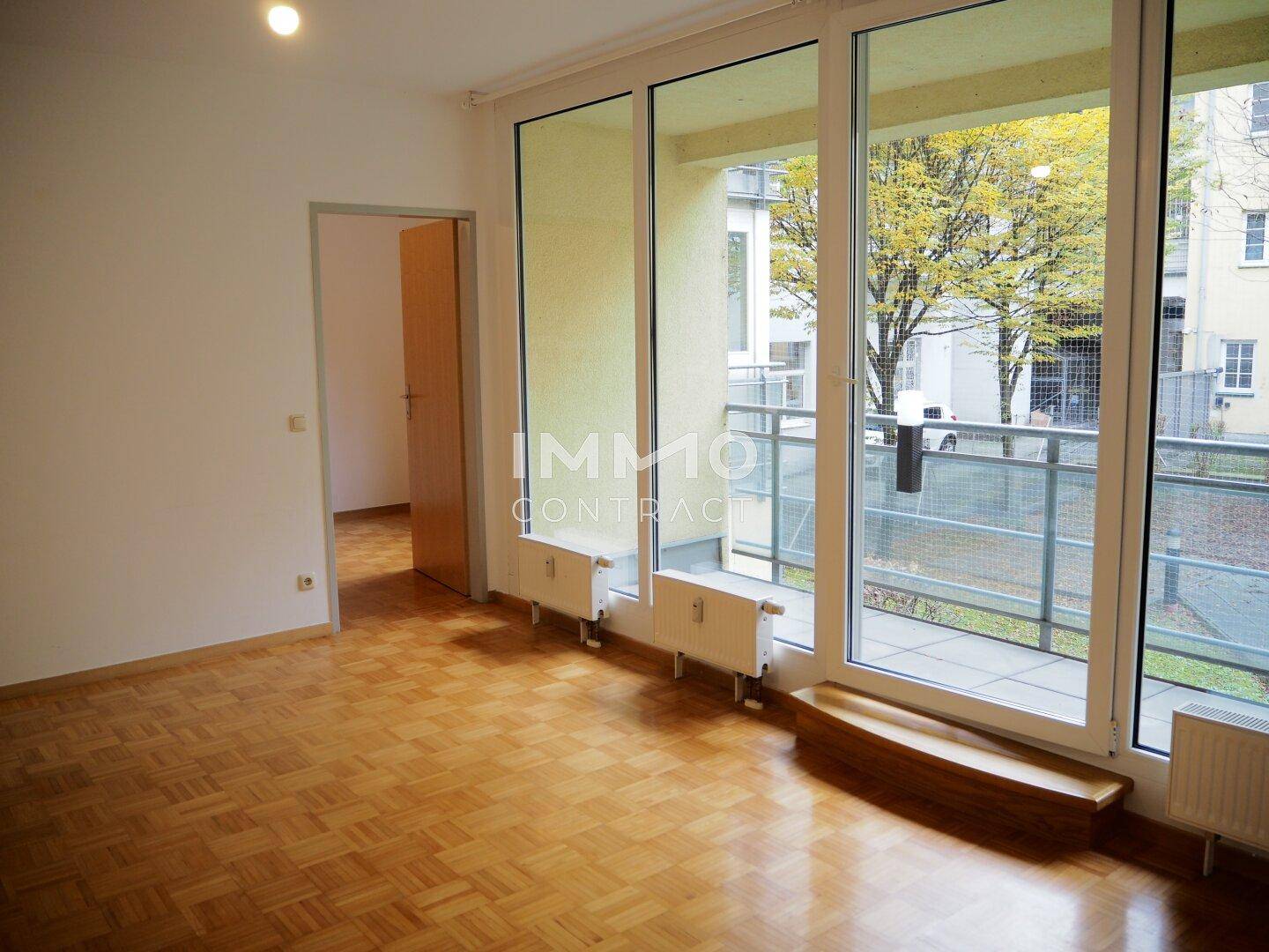 Lichtdurchflutetes Wohnzimmer mit Parkett und großen Fenstern, die den Balkon zeigen.