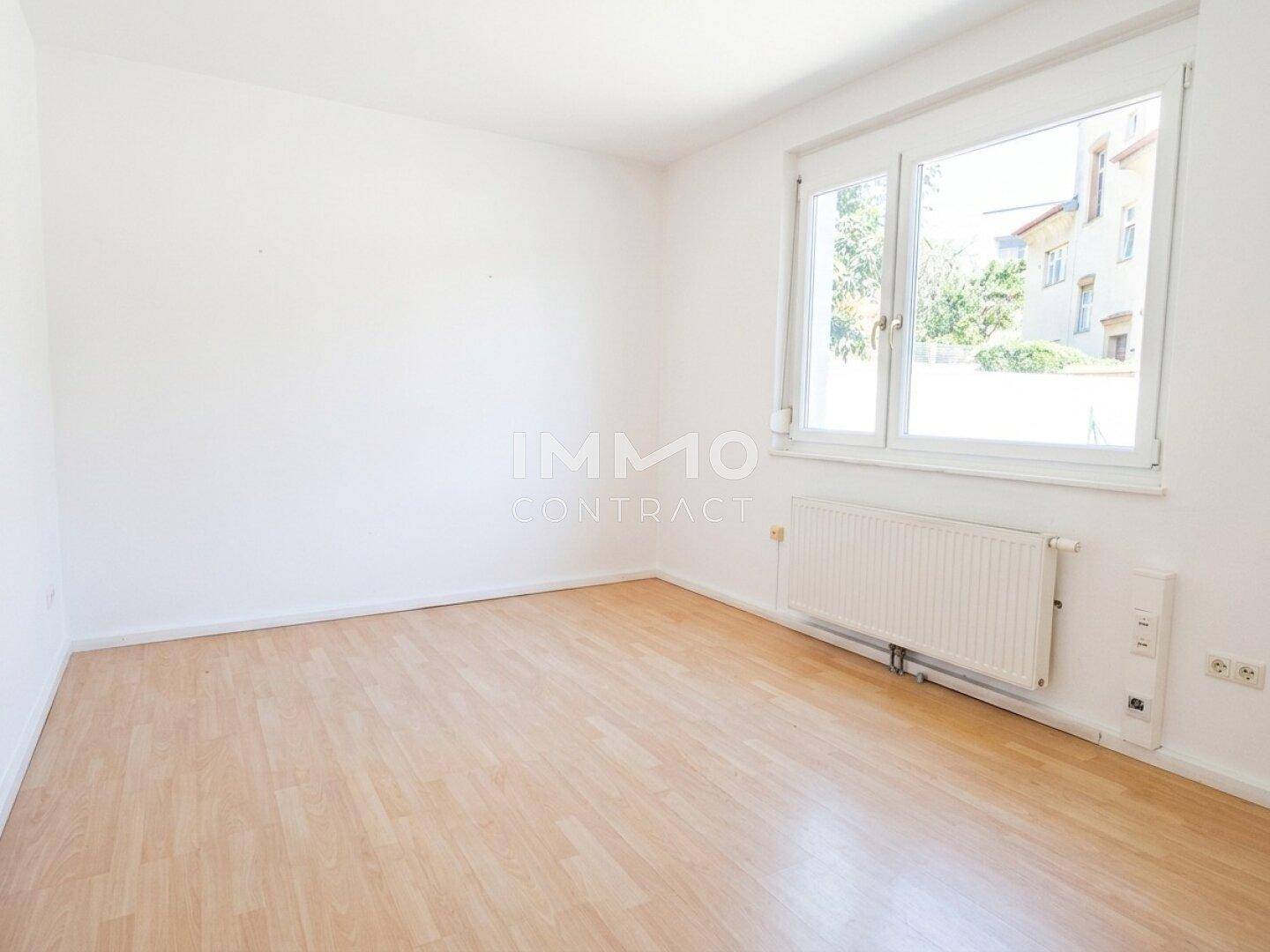 Heller, leerer Raum mit Laminatboden und großem Fenster, das viel Tageslicht hereinlässt.