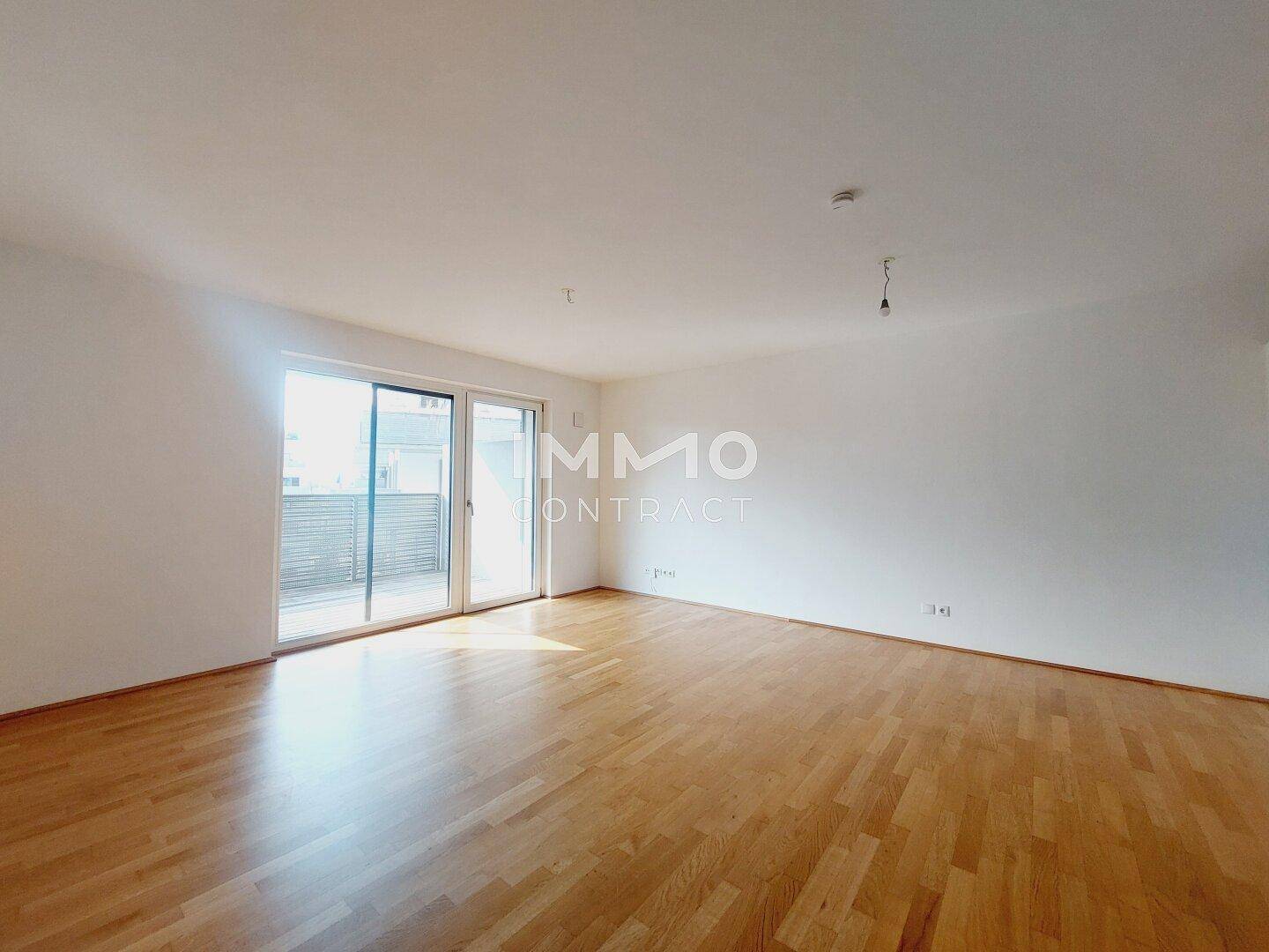 Geräumiges Wohnzimmer mit hellem Parkettboden und Zugang zu einem Balkon.
